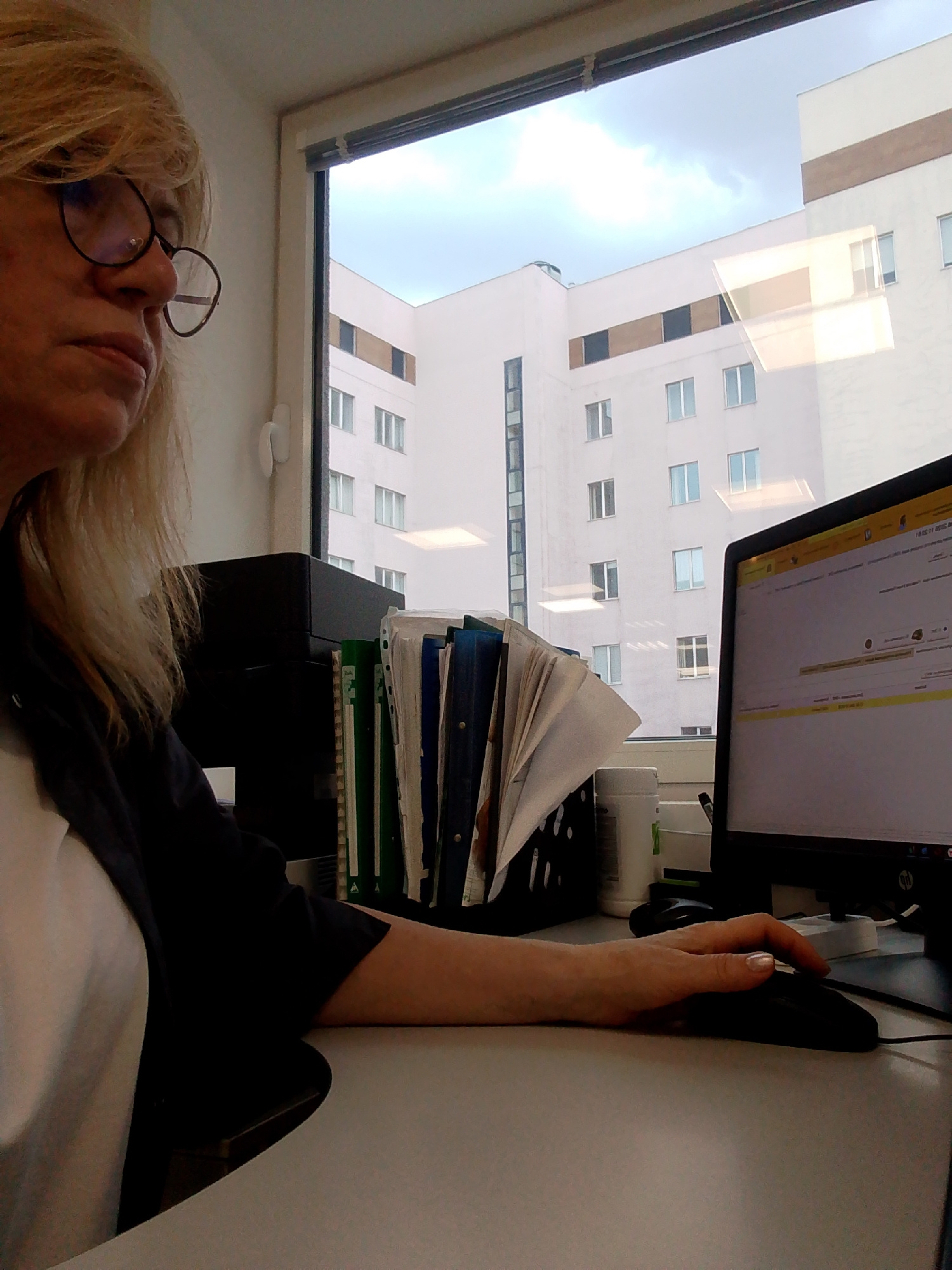 A middle-aged woman with blonde hair and glasses is seated at a desk in an office, with a computer monitor in front of her. She is wearing a black blazer over a white shirt. Her right hand is on a computer mouse, and she appears to be working. The office has a window with a view of a modern building, possibly in Moscow, Russia. There are folders and papers on the desk. The lighting suggests it is daytime, likely overcast.