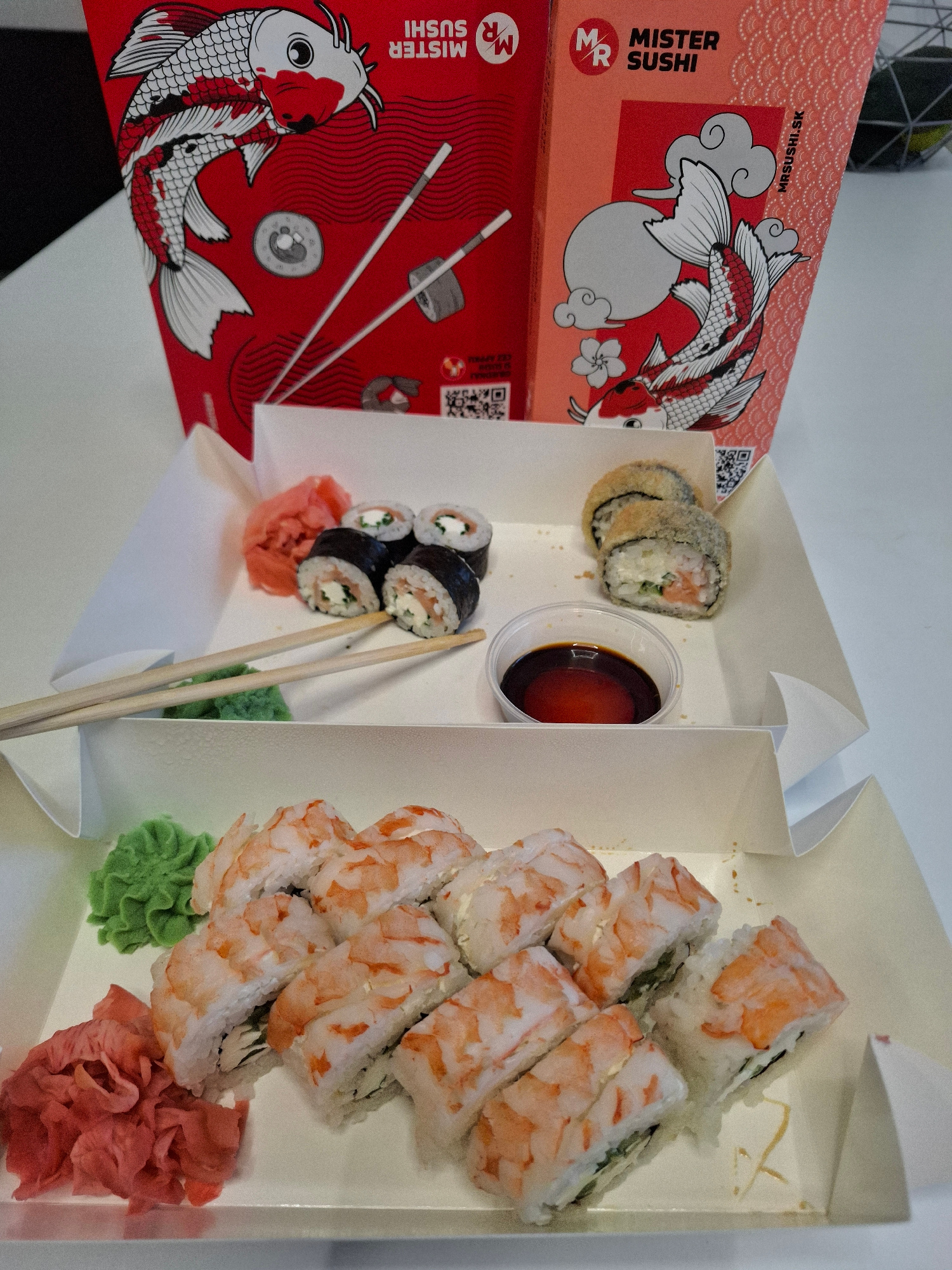 The image displays a white cardboard container holding sushi. The container is divided into two sections. The top section contains four sushi rolls, two of which appear to be fried and the other two are standard rolls with seaweed wrap, rice, salmon, and cream cheese. There is a small dish of soy sauce and a small pile of green wasabi. The bottom section of the container holds several shrimp sushi rolls. Next to the shrimp sushi rolls are slices of pickled ginger. In the background, two red cardboard boxes with a koi fish design and "MISTER SUSHI" branding are visible. The overall scene suggests a takeout sushi meal. There are no people or specific landmarks visible that would indicate the city of Bratislava, Slovakia.