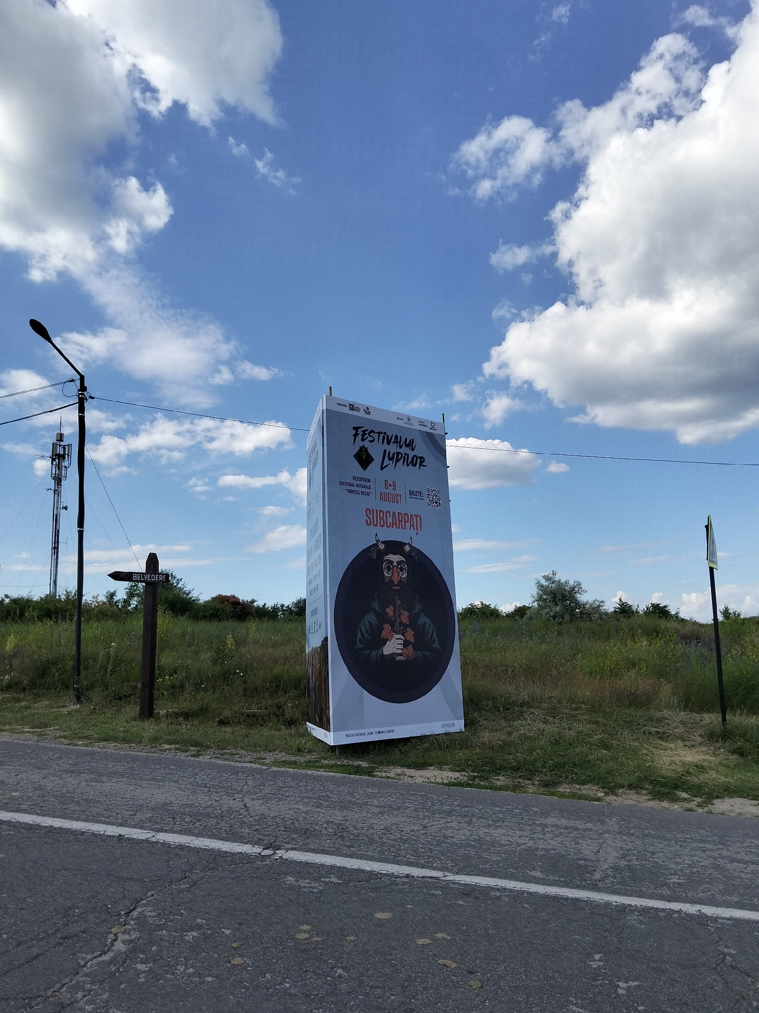 A tall, rectangular banner stands vertically on a grassy field, advertising the "FESTIVALUL LUPILOR". The banner features a dark, circular graphic of a person with horns, wearing glasses, and holding leaves. Text on the banner indicates the dates "8-9 AUGUST" and mentions "REZERVAȚIA CULTURAL-NATURALĂ 'ORHEIUL VECHI'", suggesting a cultural and natural event. The words "SUBCARPATI" and "ORIGIN" are also visible.
To the left of the banner, a wooden signpost points left with an arrow and the word "BELVEDERE" inscribed on it. In the background, a tall telecommunication tower is prominent against a bright blue sky dotted with fluffy white clouds. The scene is set by a road in the foreground, which is asphalt with a white line marking its edge. The overall impression is of an outdoor event taking place in a rural or natural setting, likely during daytime given the clear sky. The weather appears to be good, with sunshine and scattered clouds.