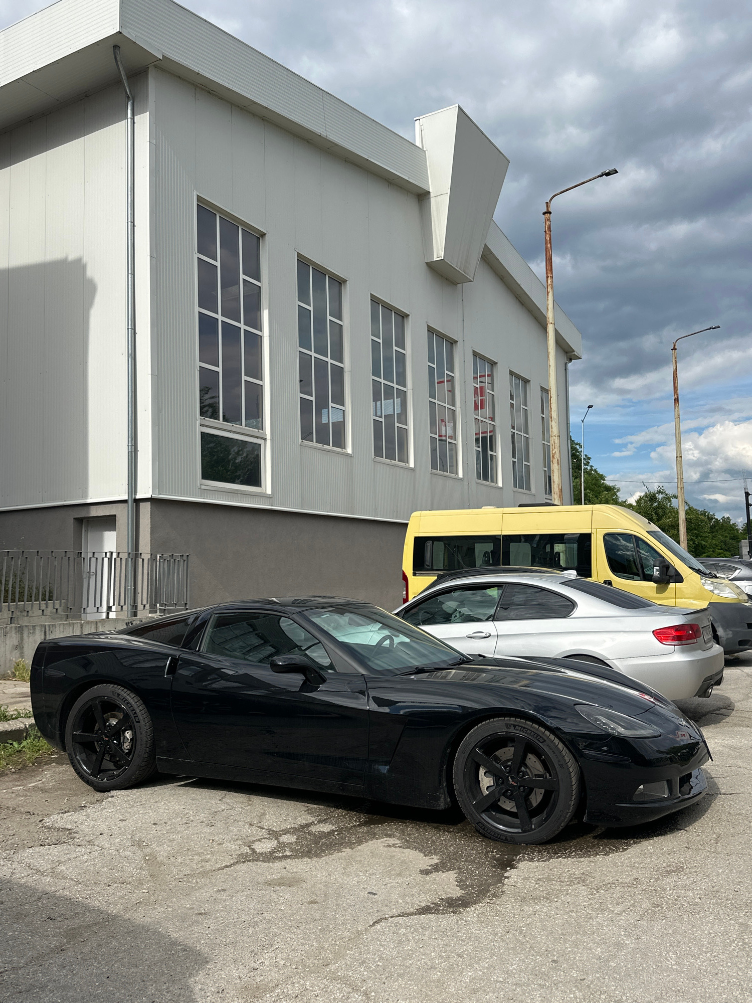 A black Chevrolet Corvette is parked on a concrete lot in front of a white industrial-style building with numerous large windows. To the right of the Corvette, a silver BMW coupe is parked, followed by a yellow van. The building has a grey base and a metal corrugated roof. Two rusty streetlights stand near the building. The sky is overcast with dramatic clouds. The image appears to have been taken in Veliko Tarnovo, Bulgaria, as suggested by a tag on an external dataset.