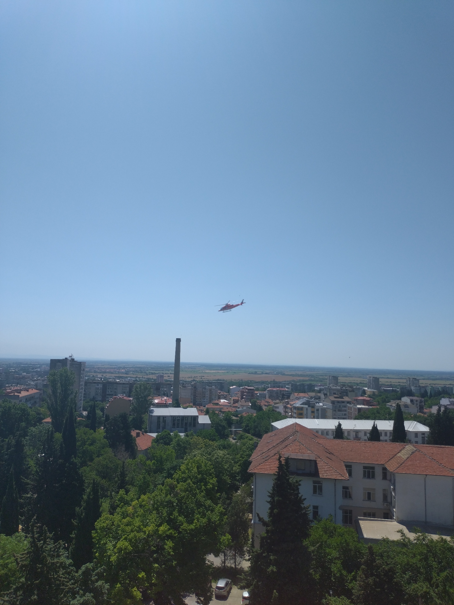The image is an aerial view of the city of Stara Zagora, Bulgaria, on a clear, sunny day. In the sky, a red helicopter is visible, flying from left to right. Below, a cityscape unfolds with a mix of residential buildings, some taller apartment blocks, and industrial structures, including a prominent tall chimney. Green trees are abundant, interspersed between the buildings and in the foreground. In the lower right corner, a white building with a red tiled roof, possibly a hospital or an institutional building, is clearly visible. The background shows a flat horizon line, suggesting the city is situated in a plain. There are no visible people or specific interactions taking place.