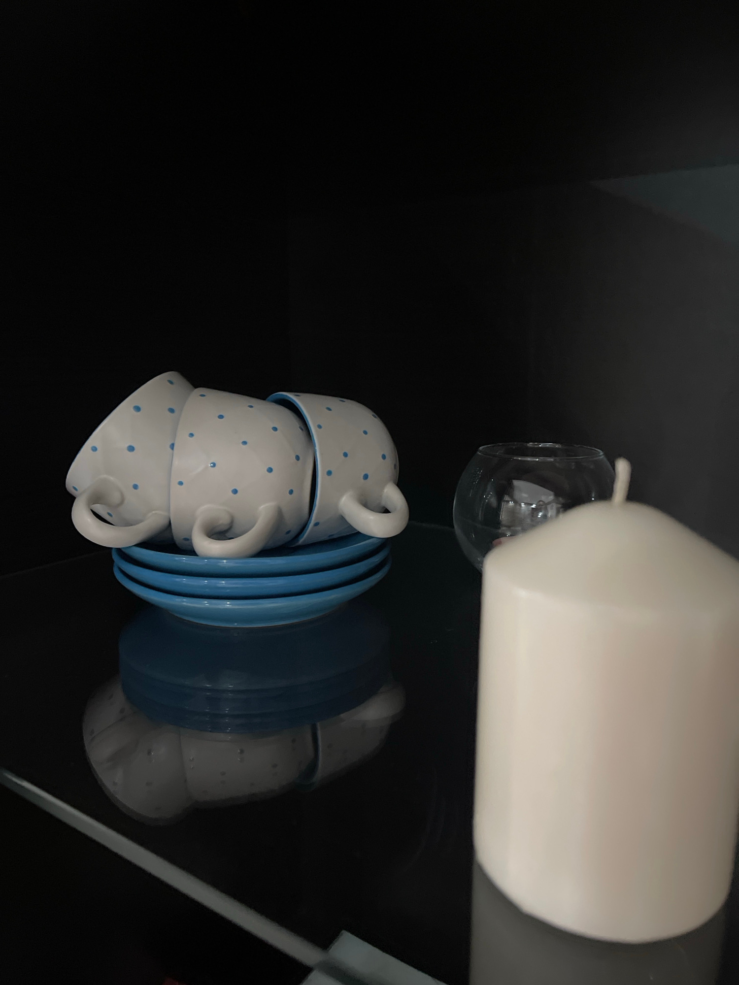 A stack of three white ceramic teacups with blue polka dots sits next to a stack of blue saucers. The cups are nested one inside the other, with their handles facing outwards. A white object, possibly a pitcher or a milk frother, is positioned above and to the right of the cups and saucers, its reflection visible in the dark, glossy surface below. The overall scene is set against a dark background, with a subtle reflection that adds depth. The lighting appears to be artificial, suggesting an indoor setting, possibly a kitchen or dining area. There are no visible people, text, or indications of weather or time of day. The location context suggests this image was taken in Stavropol, Russia, but there are no visual cues within the image to confirm this.