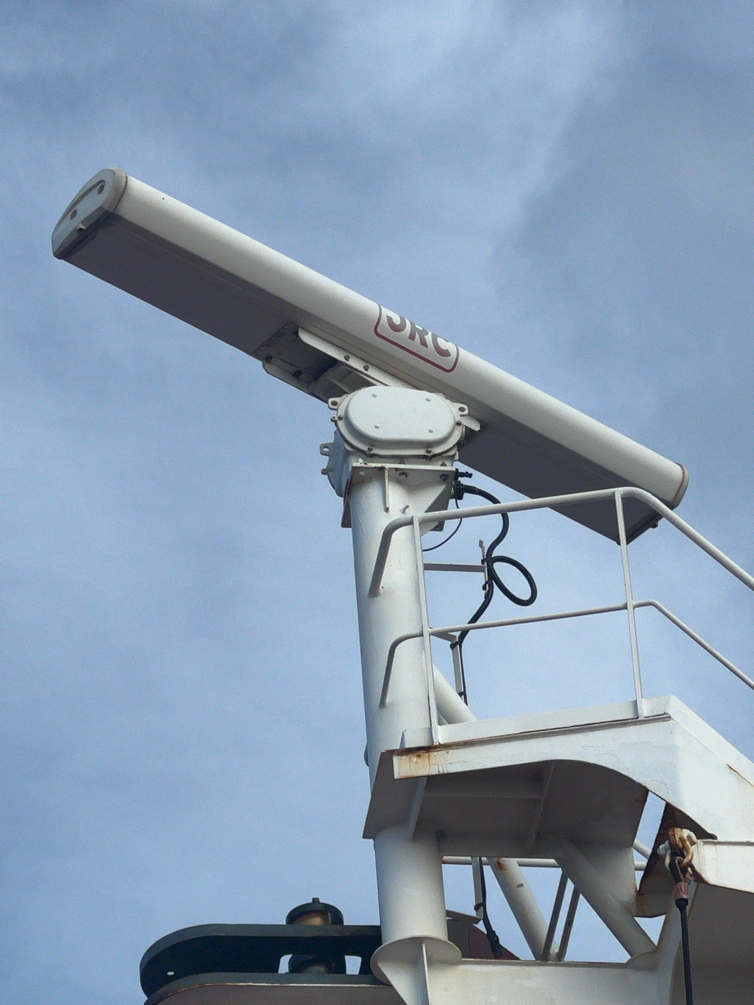 A radar unit is mounted on a white support structure against a cloudy sky. The radar unit is a long, horizontal white rectangle with a red logo that reads "JRC" on its side. The support structure is made of white painted metal, with railings and various mechanical components. The image is a close-up shot, focusing on the radar and its mounting. The location is not specified beyond "Unknown, Unknown".