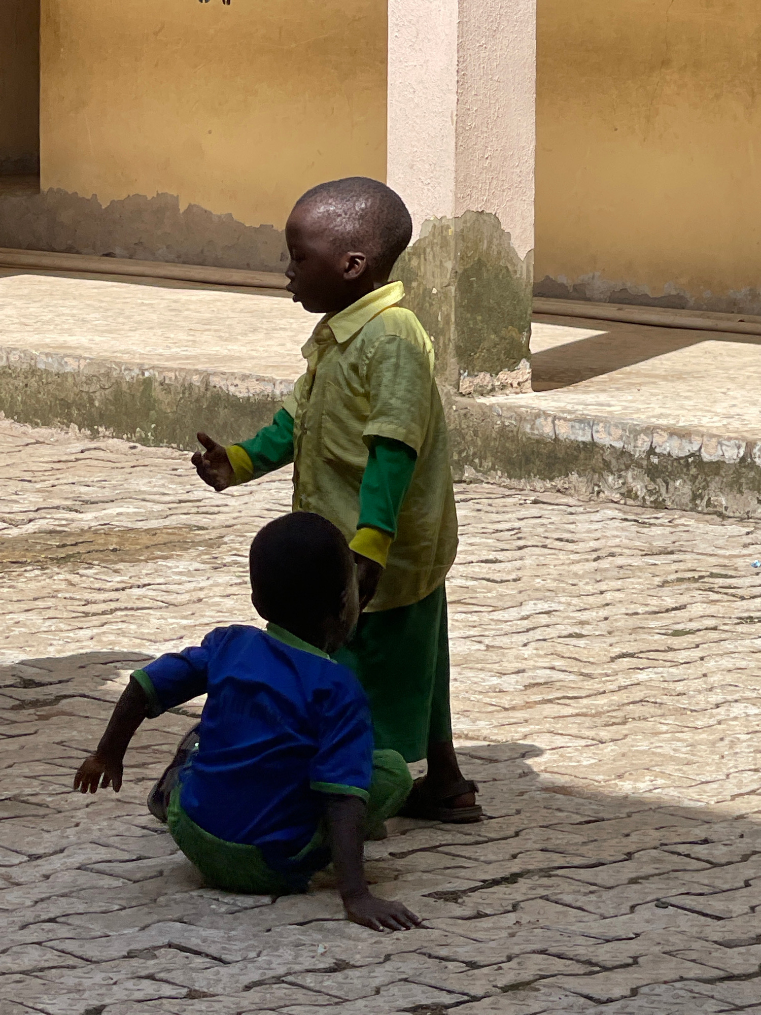 Two young boys are in an outdoor courtyard paved with interlocking bricks. The boy in the foreground is wearing a blue shirt with green trim and green pants, and he is crouched on the ground, facing away from the camera. His left arm is extended forward. The boy in the background is standing and wearing a light green short-sleeved shirt over a green long-sleeved shirt, and green pants. He is facing left and gesturing with his right hand. Behind the boys, a textured, pale yellow wall with a weathered, greenish-grey base runs along the back of the courtyard, with a white pillar to the left of it. The ground is partially shaded, indicating direct sunlight. The image appears to have been taken in Akwanga, Nigeria.