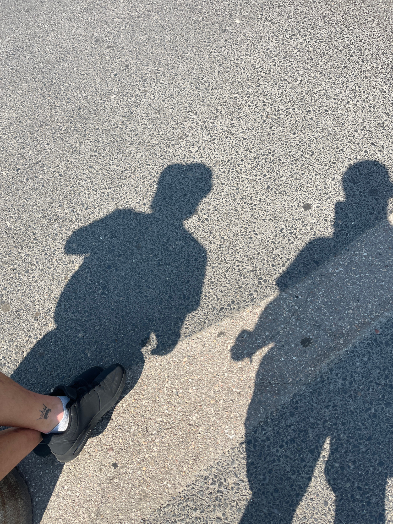 This is a high-angle, close-up shot of two shadows cast on an asphalt surface, likely a street or sidewalk in Piaseczno, Poland. The shadow on the left appears to be of a person in a squatting or sitting position, with their arm bent. The shadow on the right is of a person standing, with their arm extended. In the foreground, on the left side of the image, a person's leg and foot are visible, wearing a black sneaker and white sock. A small tattoo of a crown is visible on the person's ankle. The asphalt surface is textured and appears to be illuminated by strong sunlight, creating sharp shadows. The overall impression is one of a casual moment captured outdoors.