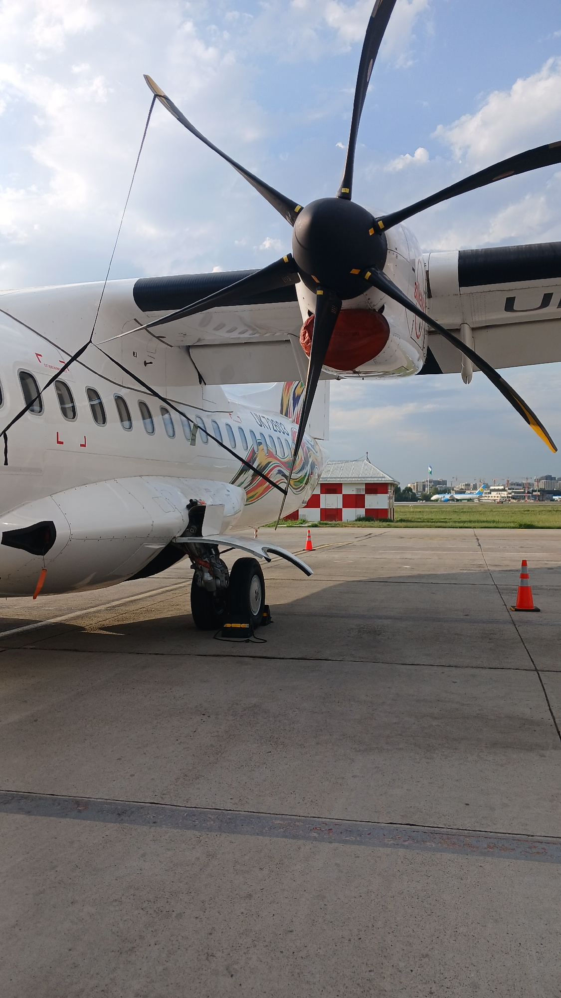 The image shows the side of a white turboprop airplane parked on an asphalt tarmac. The airplane's propeller is prominently featured in the foreground, with its black blades and yellow tips against a cloudy blue sky. Various wires are attached to the propeller and the aircraft's fuselage. The aircraft has several windows along its side, and a colorful design featuring rainbow-like streaks is visible on the tail section.
The landing gear, including a wheel and some mechanical components, is visible beneath the fuselage. In the background, there is a small building with a red and white checkered pattern, and beyond that, more airplanes are parked on the tarmac of an airport. Trees and city buildings can be seen in the far distance, suggesting an urban environment. Two orange traffic cones are placed on the tarmac.
This scene is consistent with the general appearance of the apron at Tashkent International Airport (UTC), also known as Islam Karimov Tashkent International Airport, located in Tashkent, Uzbekistan. The presence of commercial aircraft, airport infrastructure like the checkered building which could be a small control tower or utility building, and the overall layout of the tarmac are typical of such an airport.