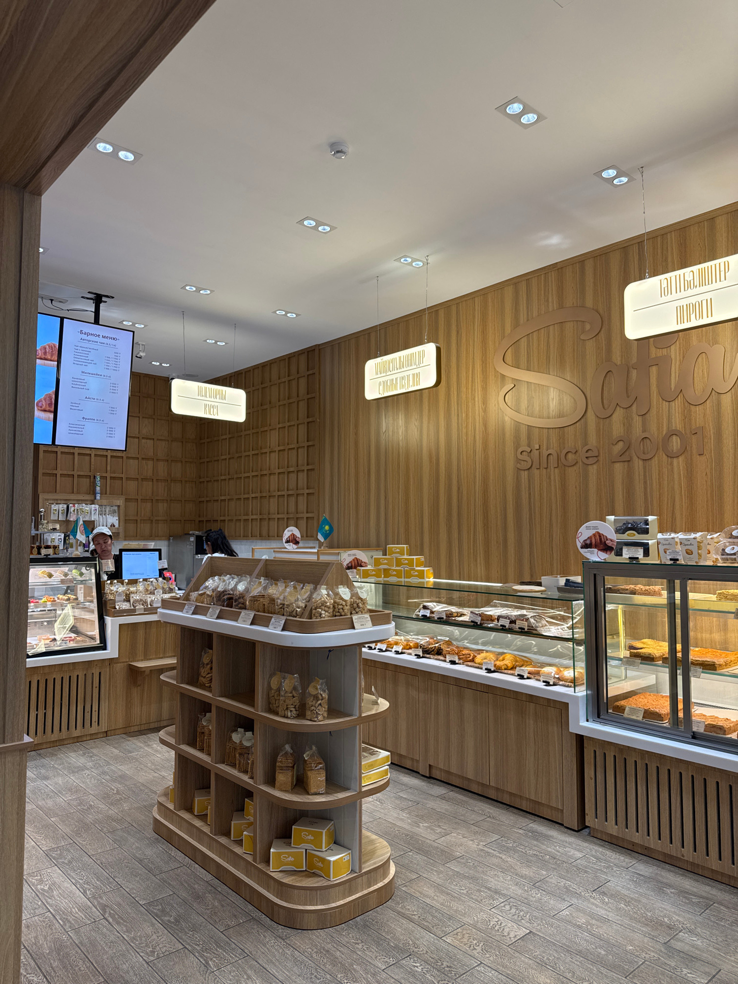 The image depicts the interior of a bakery, likely located in Almaty, Kazakhstan, as indicated by the presence of the Kazakh flag visible in the background. The bakery features a modern design with warm wood paneling on the walls and floor. In the foreground, a tiered display shelf showcases packaged snacks and boxed goods. To the left, a refrigerated display case contains various pastries and cakes. Further into the store, a counter area is visible with a computer monitor and a person attending to it. Behind the counter, another individual is partially visible. The overall atmosphere is clean and inviting, suggesting a well-maintained establishment. The branding "Sara Since 2001" is prominently displayed on the wooden wall, indicating the bakery's name and founding year. Illuminated signs are hung from the ceiling, displaying menu items in Cyrillic script.