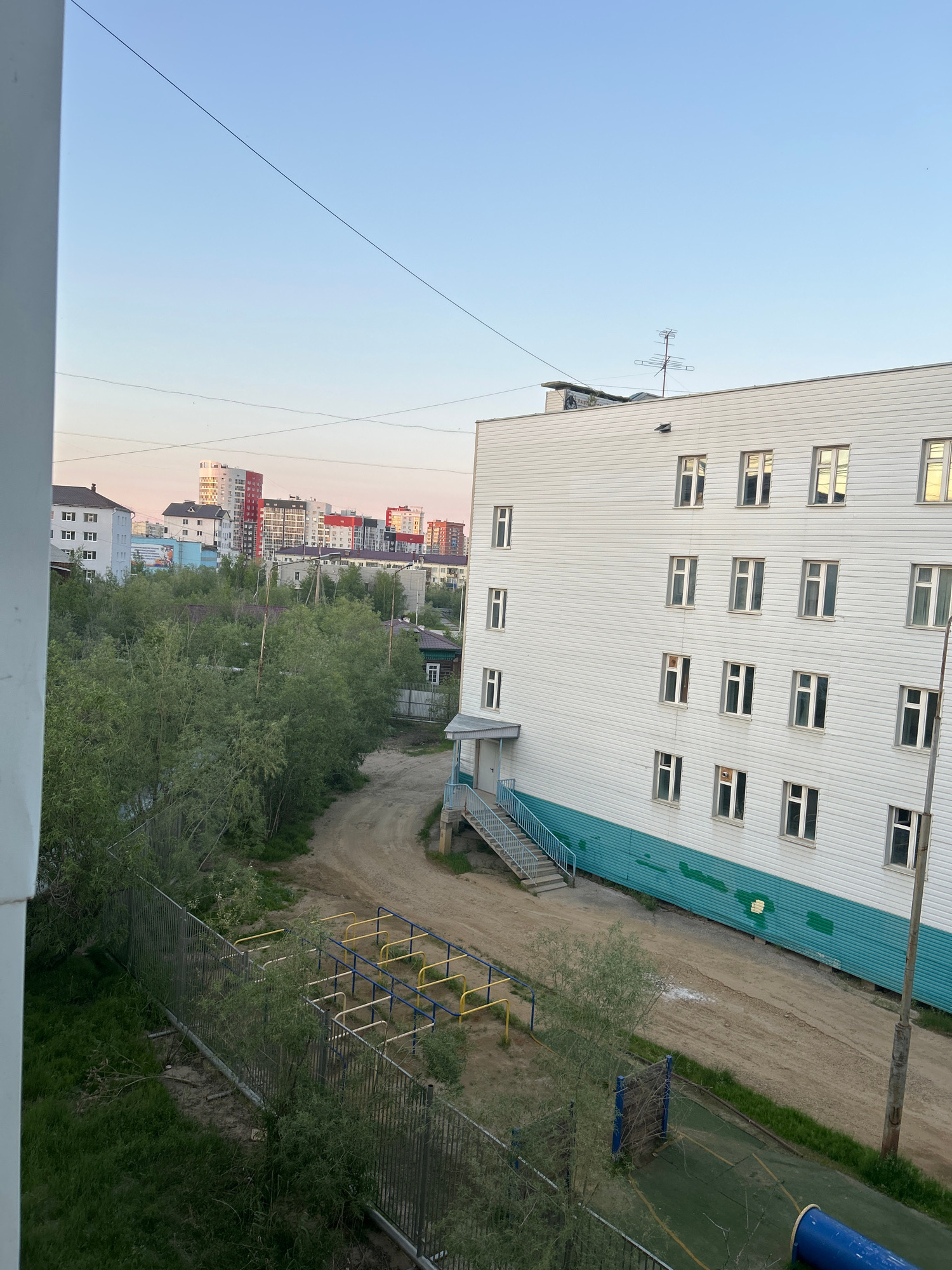 This image, taken in Yakutsk, Russia, presents an outdoor scene in what appears to be a residential area during twilight. The foreground features a section of a white building with a turquoise-green lower facade, characterized by multiple rectangular windows. To the left, a metal fence encloses an area with green grass and some vegetation. Beyond this, a dirt path winds through a sparsely treed area. Further in the background, a cluster of multi-story buildings, including some taller, modern structures with colorful facades, is visible against a pale pink and blue sky. A small playground structure with yellow and blue bars is situated near the dirt path. The overall impression is one of a suburban or outer-city environment in a northern climate, likely during the summer due to the presence of trees.
