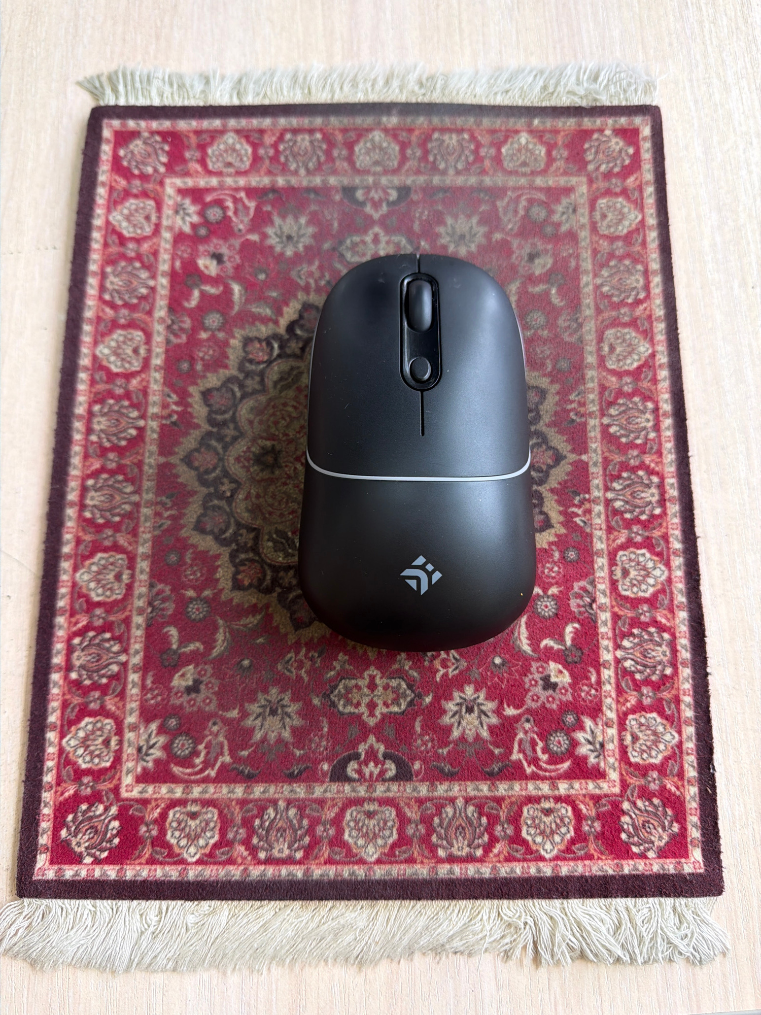A black computer mouse with a white stripe around its circumference and a small logo is positioned centrally on a miniature oriental rug with a red background and intricate gold and white patterns. The rug has a dark red border and a fringe of cream-colored threads along its bottom edge. The rug is placed on a light-colored wooden surface. The scene is viewed from directly above. The image does not provide any information about the city of Irkutsk, Russia.