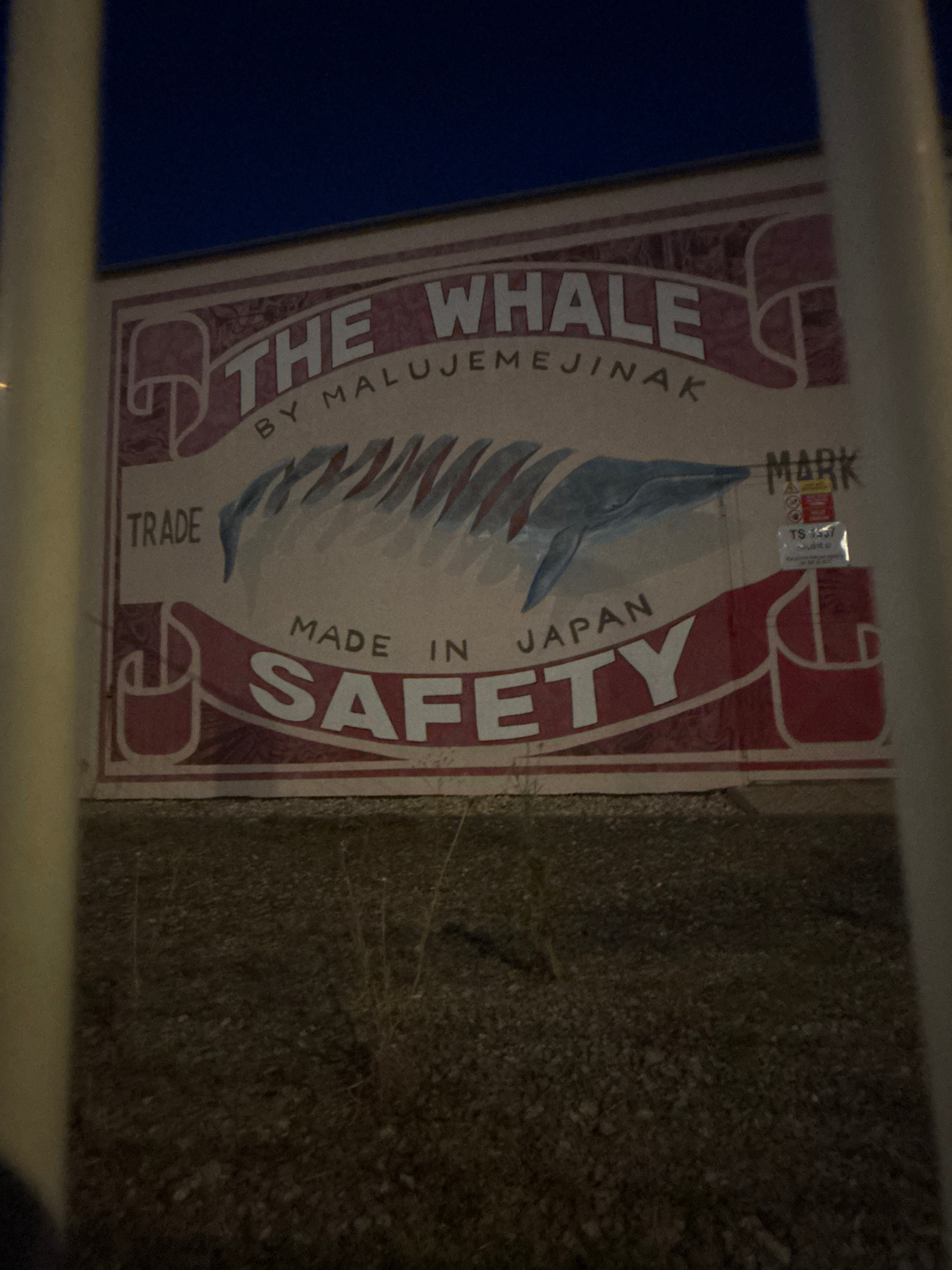 A nighttime, medium shot captures a mural painted on a light-colored wall, possibly a building exterior. The mural features a stylized design with a dark blue sky backdrop. Dominating the mural is the text "THE WHALE" in large, white, serifed letters, with "BY MALUJEMEJINAK" in smaller, dark letters below. A blue and white whale illustration is depicted in the center, with water or motion lines. To the left, "TRADE" is written in dark letters. To the right, a smaller section displays "MARK" and a label with "TS 1537". Below the whale, "MADE IN JAPAN" is written in dark letters, and "SAFETY" is in large, white, serifed letters. The mural is framed by decorative red borders and stylized shapes. The foreground shows gravel and dry vegetation. Two vertical poles, one pale yellow and one white, partially obscure the mural from view. The overall lighting suggests artificial illumination.