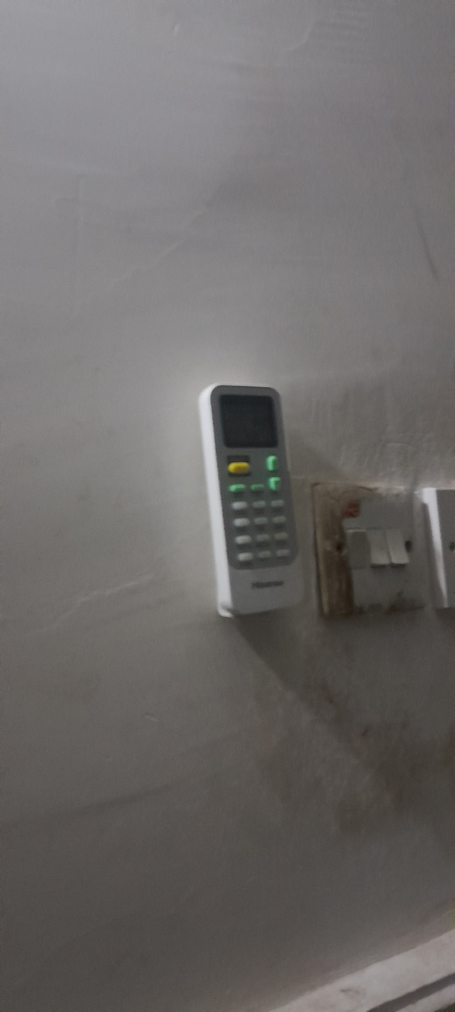 A remote control for an air conditioner is mounted on a light-colored wall, positioned vertically. The remote has a small screen at the top, a yellow button below it, and a grid of buttons underneath. Several of the buttons are illuminated with a green light. To the right of the remote, there is a light switch with two toggles. The wall appears slightly textured and shows some discoloration, suggesting it might be old or dirty. The overall lighting is dim, casting shadows on the wall. The scene depicts a typical indoor setting in an unknown location.