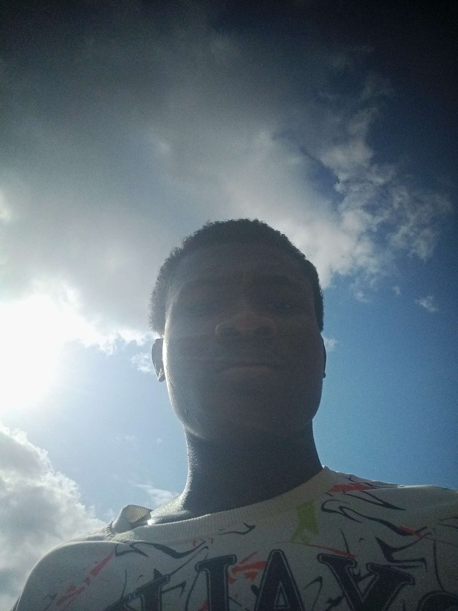 A low-angle, medium shot captures a young Black man facing forward against a bright, cloudy sky. The man's face is partially obscured by shadow due to the backlight from the sun, which is positioned to the left of the frame. He has short, dark hair and is wearing a light-colored, long-sleeved shirt with a graphic print featuring black and red abstract designs and large black letters. The background consists of a bright blue sky with scattered white clouds, some appearing dense and others wispy. The overall impression is of an outdoor setting, possibly in Fegge, Nigeria, on a sunny day with the sun positioned behind the camera or slightly to the side, creating a silhouette effect on the subject.
