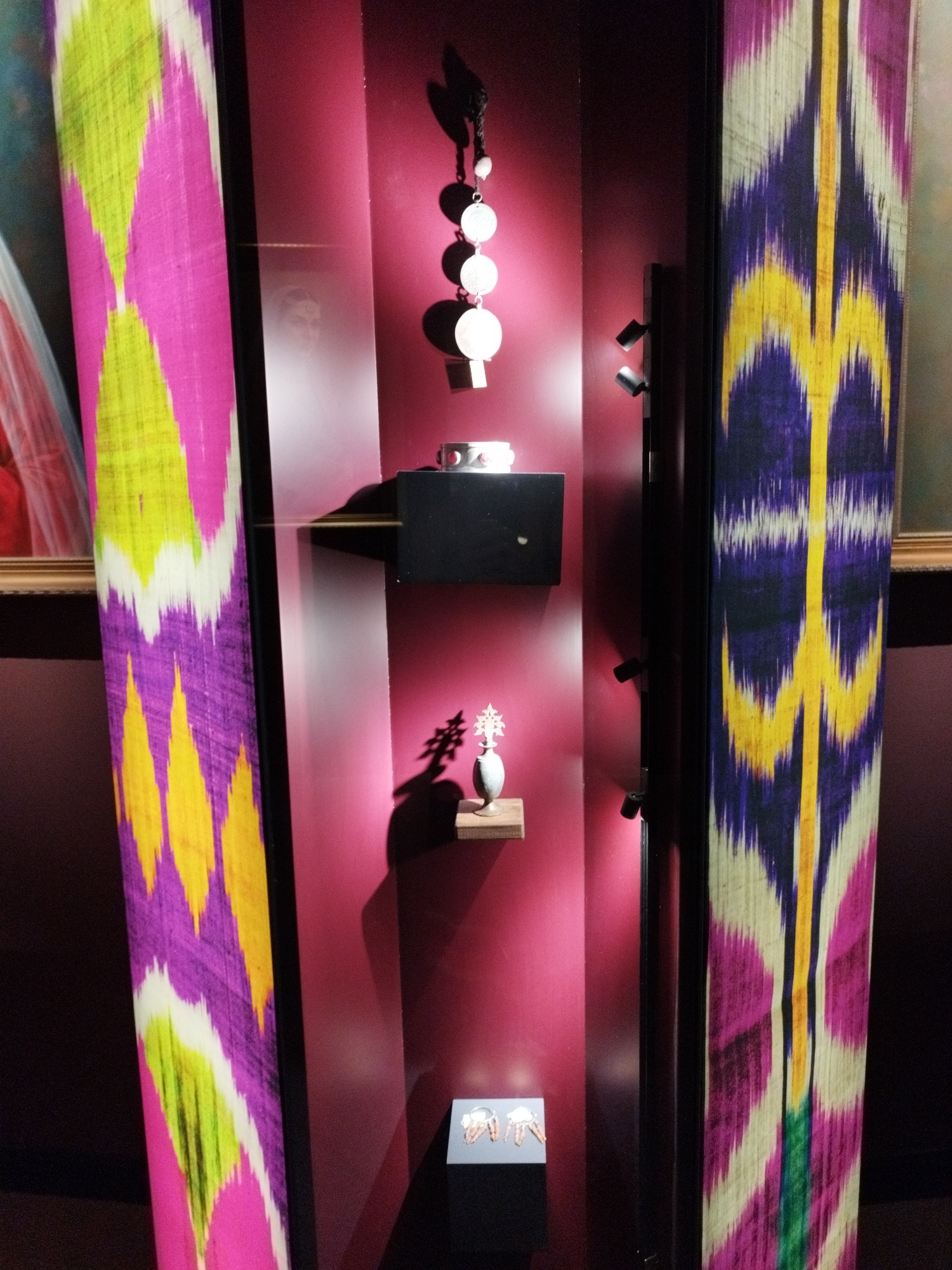The image depicts a museum display case featuring textiles with a distinctive ikat pattern in vibrant colors, including purple, yellow, green, and pink. Two tall, narrow panels of this fabric are prominently displayed on the left and right sides of the frame. Inside the display case, against a deep pink background, are several artifacts. At the top, a black cord with several round, silver-toned pendants hangs. Below this, a silver-toned bracelet or bangle rests on a black pedestal. Further down, a small, intricately shaped silver object sits atop a wooden shelf. At the bottom of the display, a small black pedestal holds two small, metallic objects that appear to be decorative charms or small sculptures, possibly representing horses. The display case is lit by spotlights, casting shadows on the wall. In the background, a hint of a painting with a golden frame is visible on the right. The overall scene suggests an exhibition of cultural artifacts or textile art.