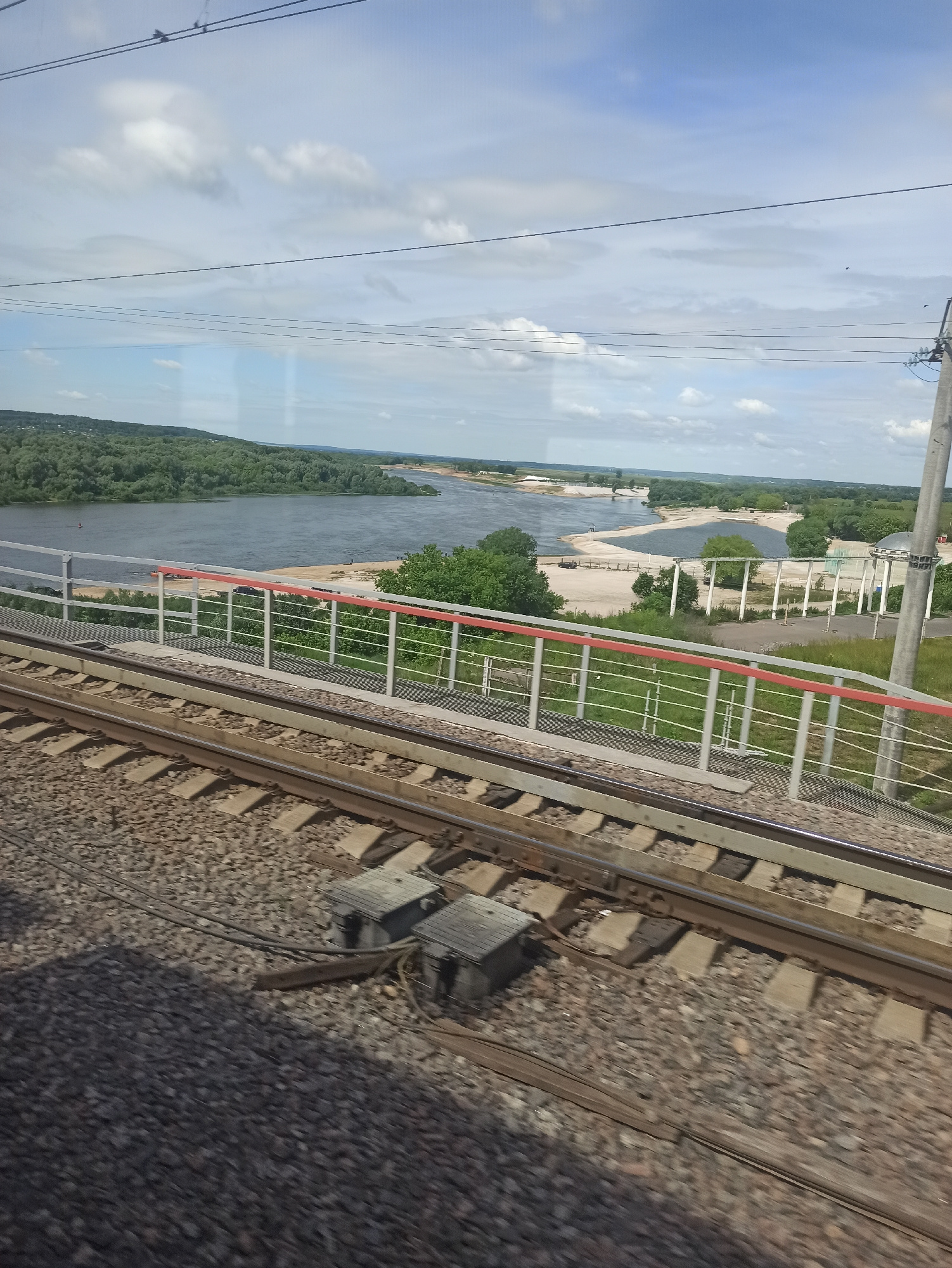 A high-angle medium shot captures a landscape viewed from a moving train. In the foreground, train tracks are laid on ballast, with two grey electrical junction boxes visible on the right side of the tracks. Beyond the tracks, a metal railing with white posts and red accents runs horizontally, separating the tracks from a beach area. A wide, calm river dominates the middle ground, with sandy shores and green trees lining its banks. In the background, rolling hills covered in lush green trees stretch towards a hazy horizon. The sky is a mix of blue with scattered white clouds. The scene suggests a serene natural environment, possibly a riverfront park or a recreational area accessible by train.
