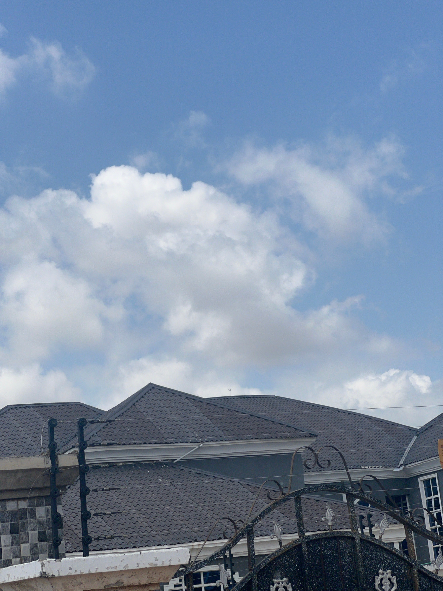 The image shows the exterior of a modern building with a dark, textured roof and decorative metal railings. The building has several windows visible, with white frames. The sky is blue with scattered white clouds, suggesting daytime. The photo is taken from a low angle, looking upwards. There is no visible text in the image. The scene appears to be in Howayi, Nigeria, indicating an urban or suburban setting. The composition focuses on the architectural details of the building against the backdrop of the sky. The lighting and clear sky suggest fair weather conditions.