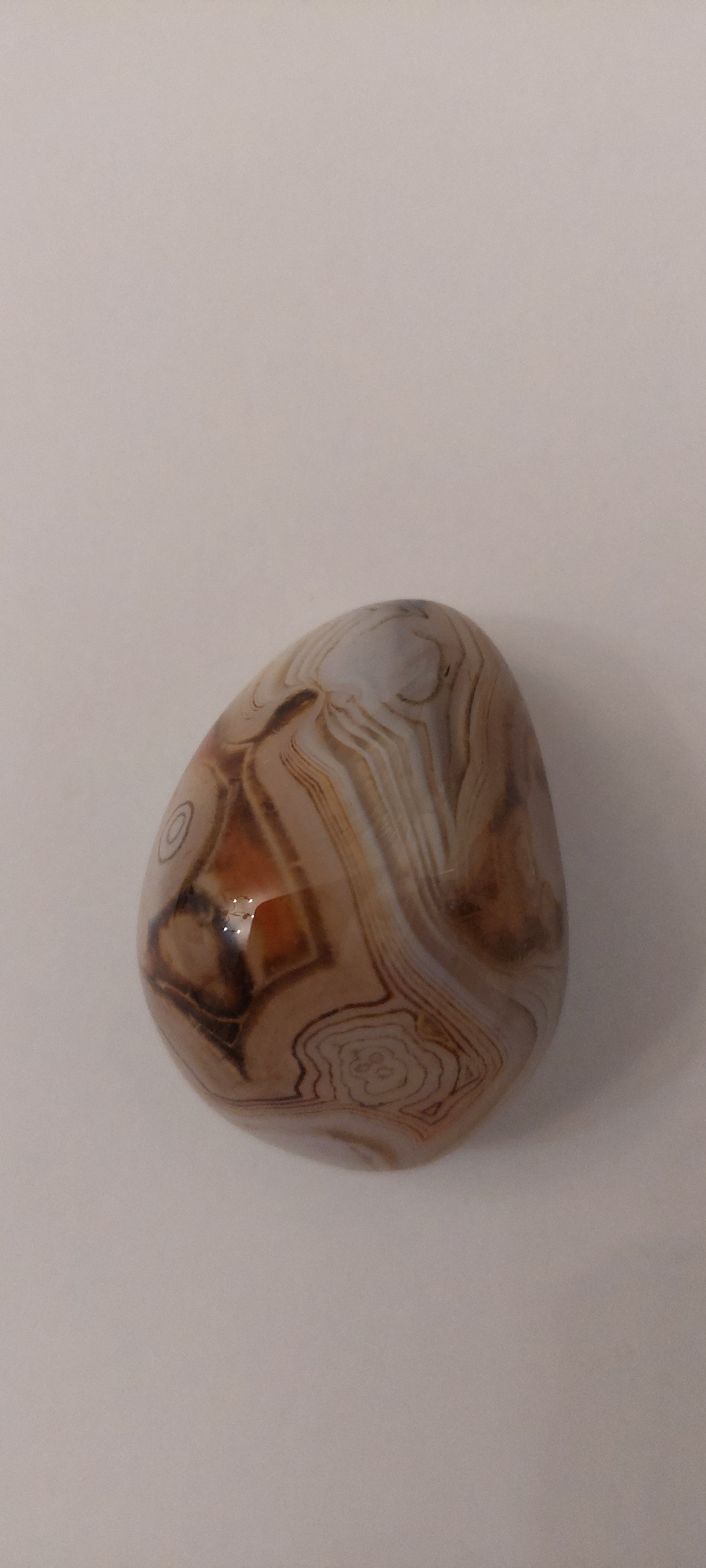 A polished, irregularly shaped agate stone lies on a plain, light-colored background. The stone exhibits intricate banding patterns in shades of brown, white, and tan, with some areas of reddish-brown. The surface of the stone is smooth and slightly reflective, showing a subtle sheen. The background appears to be a solid, neutral surface, possibly a table or paper, with soft, diffused lighting. There is a faint shadow cast by the stone on the lower right side, indicating the direction of the light source. The scene is static and the stone is the sole subject. The image does not depict any urban environments or specific locations within Ivanovo, Russia; it is a close-up studio shot of the agate stone.