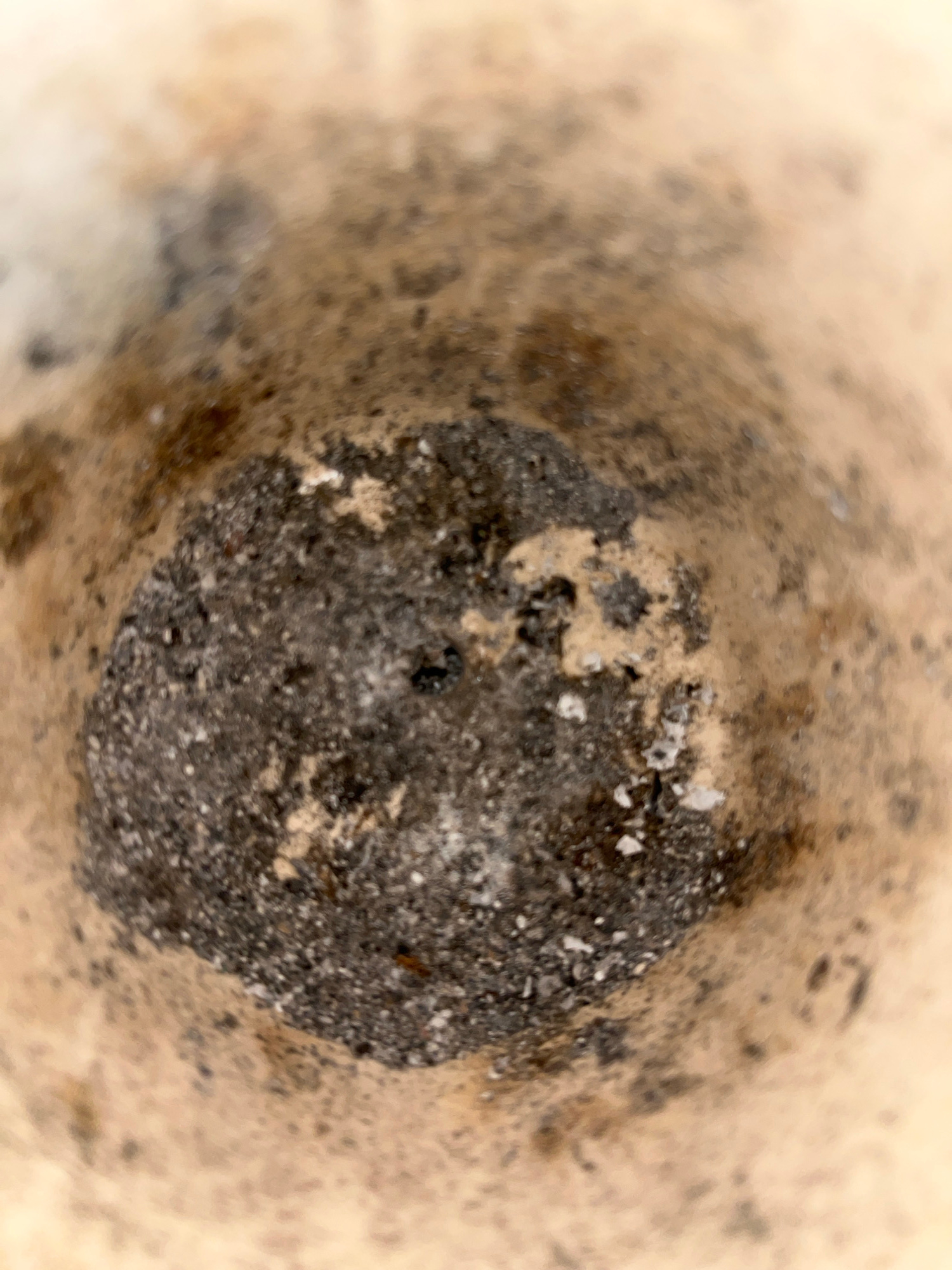 The image depicts a close-up, blurred view looking down into a rough, conical container, possibly a mortar or a waste receptacle. The interior walls are a pale, speckled brown, transitioning to a darker, more granular material at the bottom. This bottom material is predominantly dark gray to black, with visible lighter specks and irregular textures, suggesting a mixture of ash, debris, or coarse powder. The lighting appears to be overhead and diffused, casting soft shadows. There are no visible people, animals, or clear signs of specific activity. The context of "Kota Kuala Simpang, Indonesia" is not visually derivable from the image itself and would require external information.