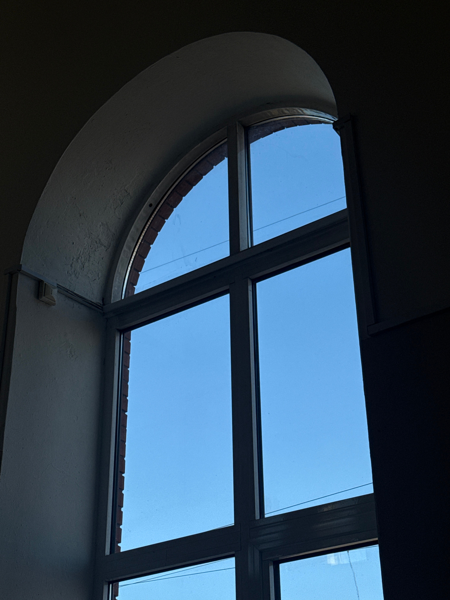 An interior shot captures a large, arched window with multiple panes against a clear blue sky. The window frame is dark, contrasting with the bright sky. The left side of the frame shows a textured, light-colored wall with a small white electrical box mounted on it. The right side of the frame is in shadow, obscuring the details of that area. The brickwork surrounding the upper arched part of the window is visible. The overall impression is of a room looking out onto a bright day through a substantial architectural feature.