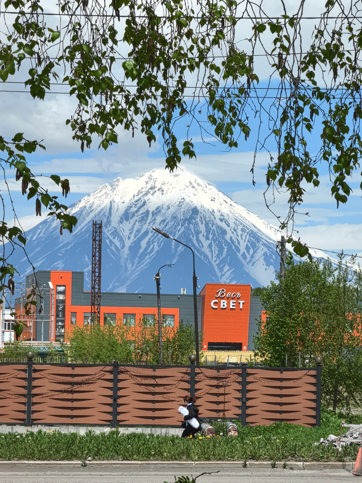 In this outdoor, daytime scene, a snow-capped mountain range dominates the background. In the mid-ground, an orange and gray building with the Cyrillic text "Весо СВЕТ" is visible. A brown, decorative fence with a woven pattern runs across the foreground. To the right of the fence, a person in black and white clothing walks to the left, carrying a white cylindrical object. Several other objects, including what appear to be rolled-up mats and construction debris, are scattered on the ground near the fence and the person. Green foliage, including leaves hanging from branches and bushes, frames the scene. The sky is blue with scattered white clouds.