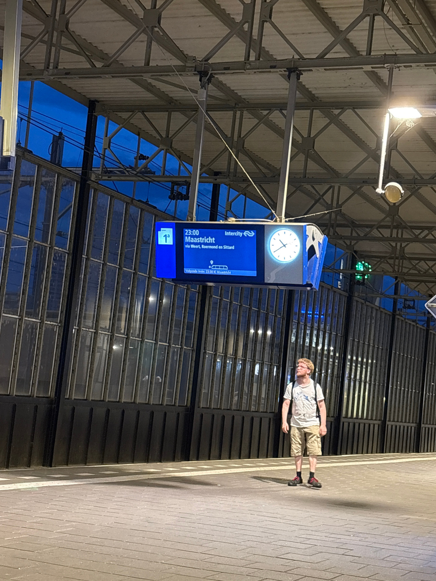 The image depicts a man standing on a train station platform in Eindhoven, Netherlands. He is looking upwards towards a digital train information board. The board shows that a train is departing at 23:00 for Maastricht, with stops in Weert, Roermond, and Sittard. The man is wearing a light-colored t-shirt, khaki shorts, and black shoes with red accents. He is carrying a backpack. The platform is paved with brick-like tiles and has white markings. Behind the platform is a dark, modern facade with vertical window panes. Overhead, the station's ceiling structure is visible, along with lighting fixtures and the train information board, which also displays an analog clock. The sky is dark blue, indicating it is either early morning or evening.