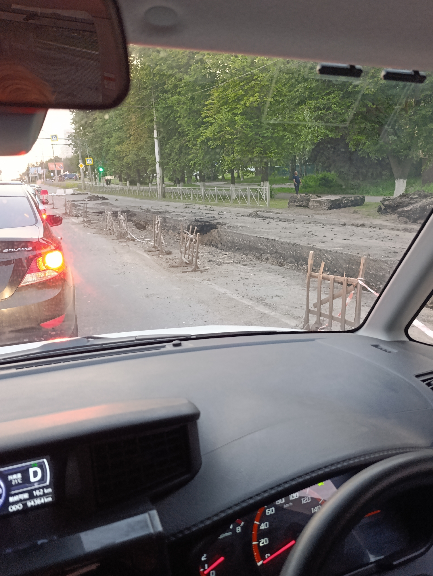 The image shows a view from inside a car, looking forward through the windshield and at the dashboard. The car is stopped in traffic on a street that is under construction. The road ahead is torn up, with trenches and piles of dirt and debris. Wooden barriers and caution tape are used to cordon off the construction area. A black car with its brake lights on is visible in front of the camera car. On the right side of the road, there are trees and a white fence. A traffic light showing green is visible in the distance, along with a sign that reads "СТОП" (STOP). A person in a dark shirt and pants is standing near the construction debris on the right side of the road. The car's dashboard displays the gear selector in "D" (Drive), the outside temperature at 21 degrees Celsius, a distance of 162 km, and the odometer reading 94364 km. The speedometer shows a speed of approximately 0 km/h. The lighting suggests it is either early morning or late afternoon. The presence of the "СТОП" sign and the general appearance of the street with trees and a fence are consistent with residential or suburban areas in Penza, Russia.
