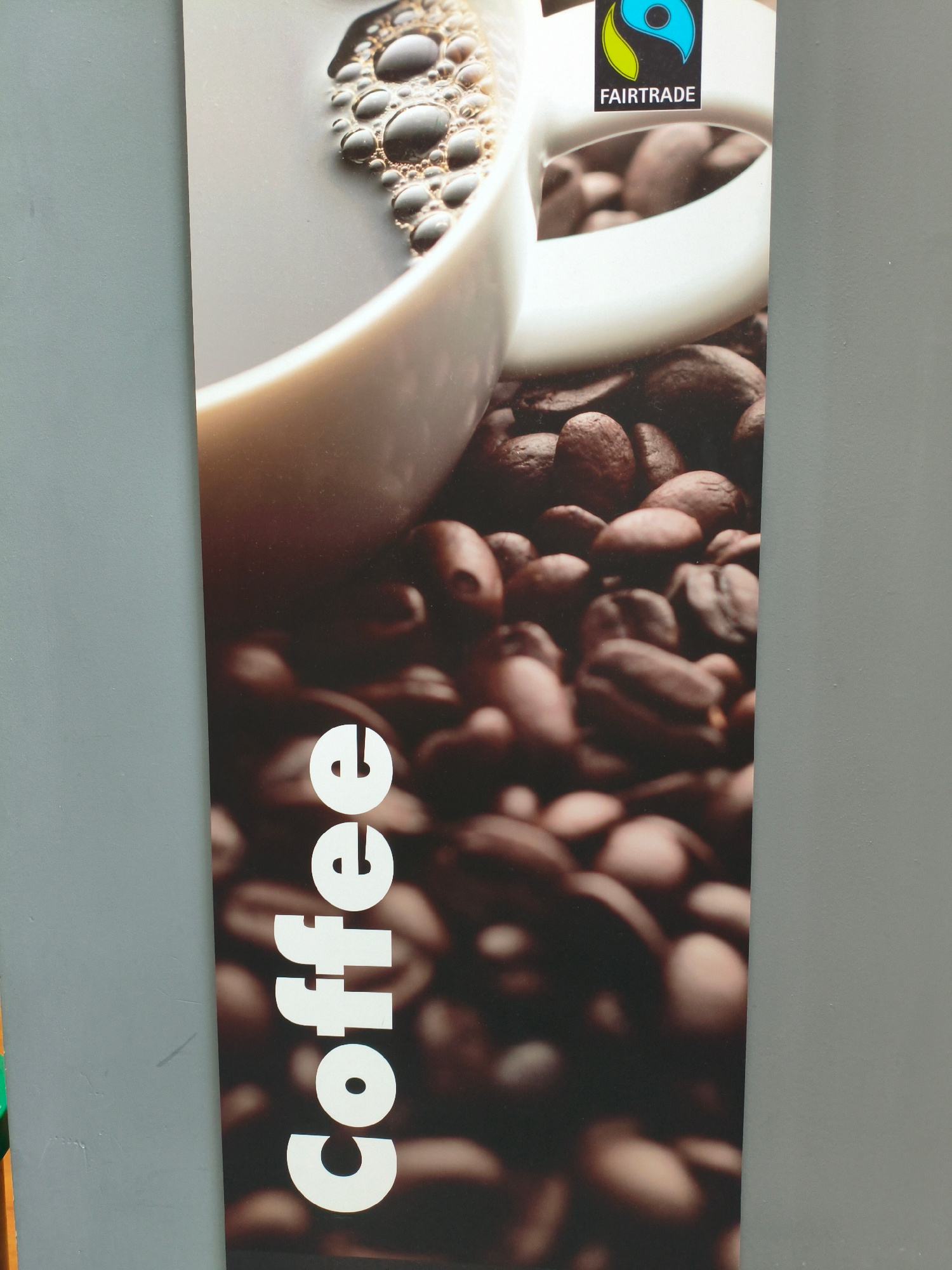 This media file is a vertical, close-up photograph of a poster or advertisement. The poster features a large, out-of-focus image of coffee beans, with a white coffee cup and some coffee with foam visible at the top. A Fairtrade logo is also present on the cup. The word "Coffee" is printed vertically in large, white, sans-serif font along the left side of the poster. The background of the image is a plain, textured gray wall, suggesting the poster is displayed indoors, possibly in a shop or cafe. The lighting appears to be even, indicating artificial light. There is no indication of time of day, weather, or specific emotions. The only text visible is the word "Coffee" and the "FAIRTRADE" logo.