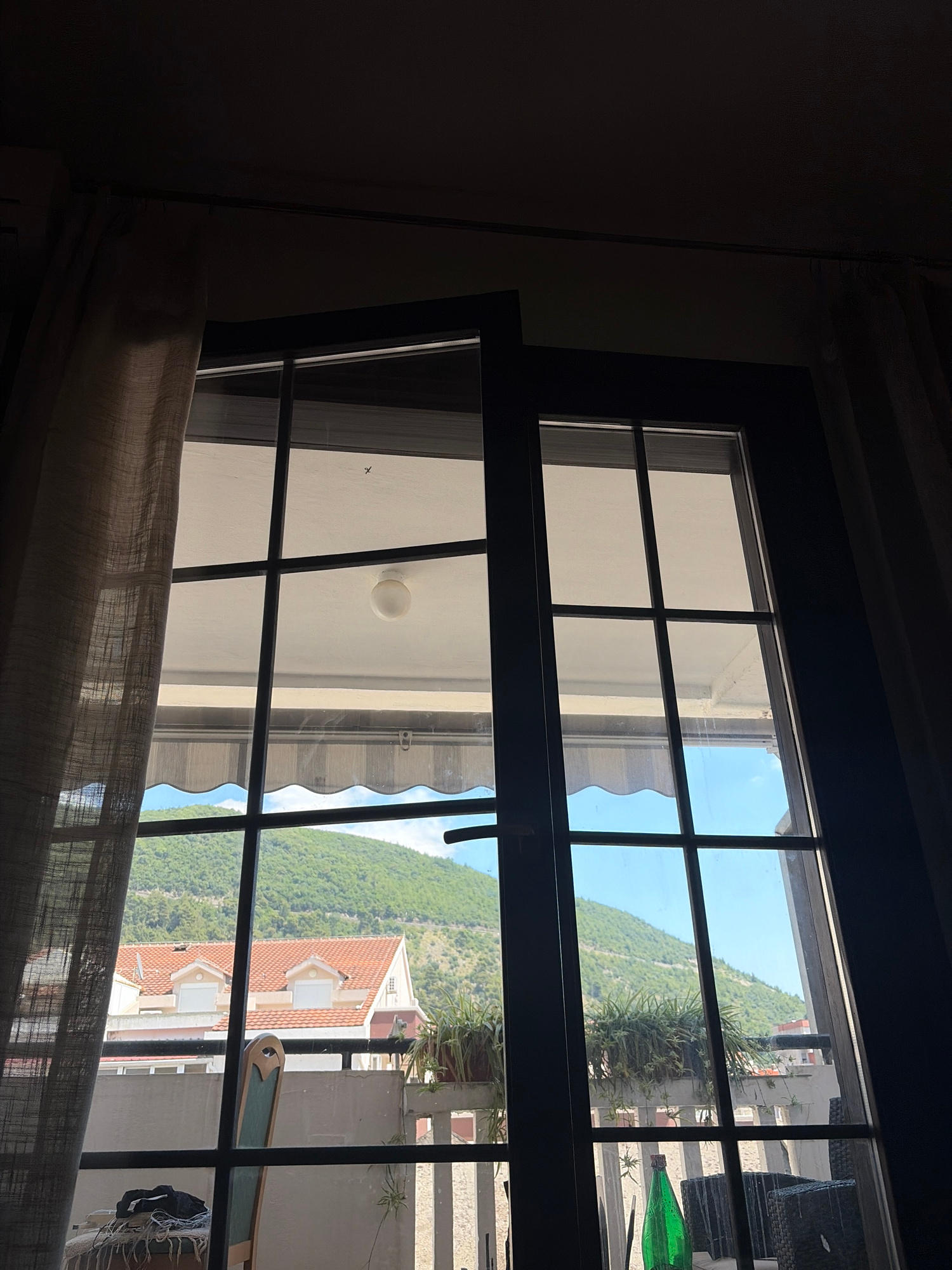 The image shows a view through a window with a grid pattern onto a balcony and beyond to a hillside with buildings and vegetation. The window has dark-colored frames. On the left, a sheer, beige curtain is partially drawn.
The balcony is visible through the lower panes of the window. There are planters with green foliage, one prominently in the upper right quadrant. To the left, a chair with a light green upholstered seat and wooden frame is partially visible. On the balcony surface, a dark bag with white cords is on top of a woven mat or blanket. A green glass bottle stands upright on the balcony floor in the lower right portion of the frame. Outdoor furniture, possibly a dark woven chair or table, is also visible to the right.
Beyond the balcony, red-tiled roofs of buildings are visible on the lower slopes of a verdant, green hillside. The sky is a clear, bright blue.
The scene appears to be a residential setting, likely an apartment or house with a balcony, in a city like Budva, Montenegro, given the architecture of the buildings and the lush hillside characteristic of the region. The lighting suggests it is daytime, with sunlight illuminating the outdoor scene.