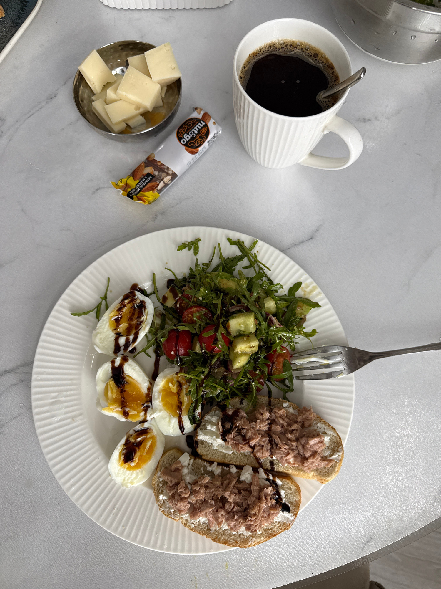A high-angle, medium shot captures a breakfast spread on a white, marble-patterned table. In the upper left, a small, silver, textured bowl contains several cubes of light yellow cheese. Next to the bowl, a white and gold wrapper with the word "nutrigoo" is partially visible. In the upper right, a white ceramic mug with vertical ridges is filled with dark coffee, and a silver spoon rests inside. The main subject is a white, fluted plate in the center of the frame, laden with food. Three halved hard-boiled eggs, drizzled with a dark sauce, are positioned on the left side of the plate. To their right, a salad composed of mixed greens, cherry tomatoes, diced cucumber, and possibly avocado is visible. In the lower right portion of the plate are two open-faced sandwiches, each topped with a white spread, likely cream cheese, and shredded tuna. A fork rests on the edge of the plate. The background consists of the white, marble-patterned table. The scene depicts a breakfast setting, likely in a home environment in Nizhnyayarkeyevo, Russia, given the context. There are no people visible in the image.