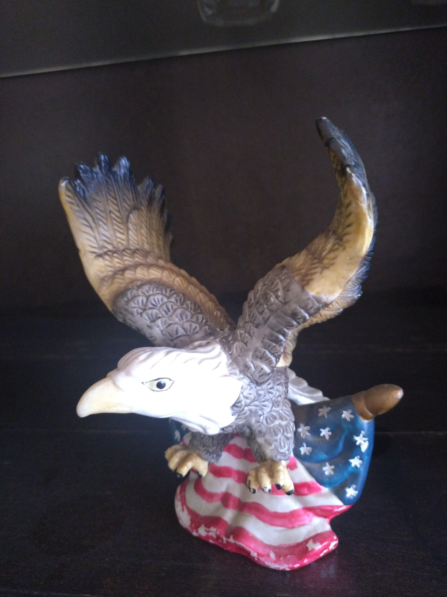A ceramic figurine of an eagle is displayed on a dark surface. The eagle is depicted with its wings spread and is perched on an American flag. The figurine is detailed with feathers and has a white head with a yellow beak and black eyes. The flag shows red and white stripes and blue with white stars. The background is dark and out of focus, suggesting an indoor setting. There is no indication of a specific location in Stara Zagora, Bulgaria.