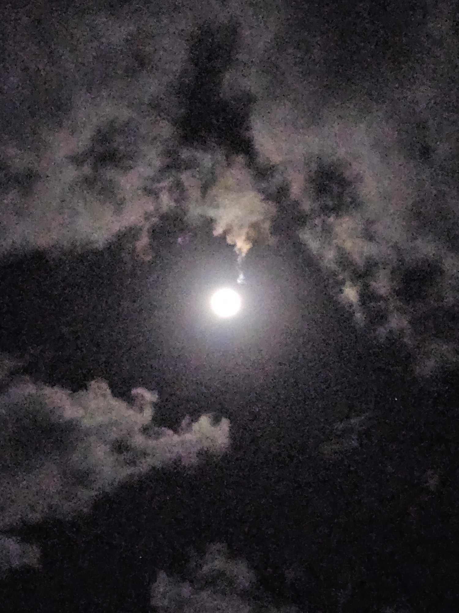 A full moon is visible in the night sky, partially obscured by clouds. The moon is bright and appears as a white circle. The clouds are dark and wispy, with some areas illuminated by the moonlight. The image was taken in Denpasar, Indonesia.