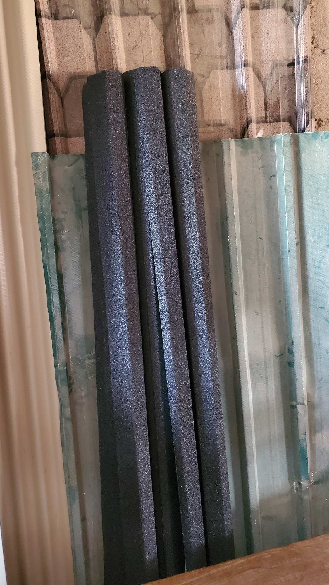 The image shows three dark gray, textured foam-like cylindrical objects stacked vertically. They are leaning against a corrugated translucent green sheet. Behind the green sheet, there is a light beige corrugated surface. To the left of the foam objects and the green sheet, a beige corrugated surface is visible. The scene appears to be outdoors, possibly in a storage area or construction site. There is no indication of Jalingo, Nigeria, or any specific location within a city. The image is a still photograph with no actions or interactions depicted.