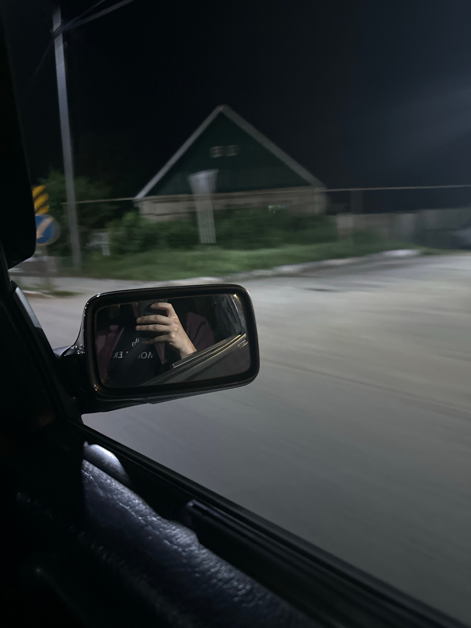 The image captures a night-time view from inside a moving vehicle, looking towards the right side mirror. The side mirror reflects a person's hand holding a smartphone, with a portion of their maroon shirt visible. The background visible through the side window is blurred due to motion, showing a road and vegetation. In the far distance, a green house with a triangular roof is discernible. The overall scene suggests a drive through a suburban or rural area at night. The location is specified as the city of Kostanay, Kazakhstan.