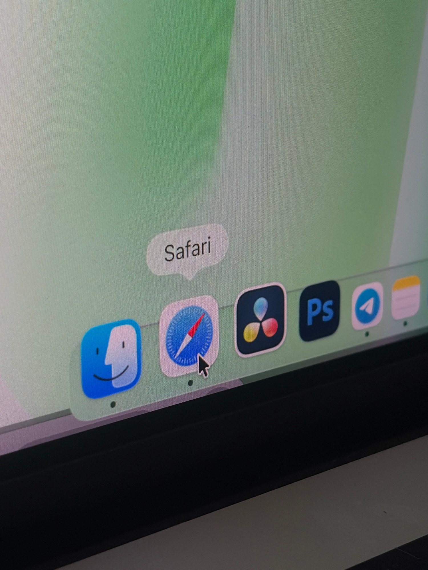 A screenshot shows a computer screen displaying a dock with several application icons, including Finder, Safari, DaVinci Resolve, Photoshop, Telegram, and Notes. A speech bubble above the Safari icon reads "Safari". The background of the screen is a gradient of light green. The lower portion of the image shows the black bezel of a laptop.