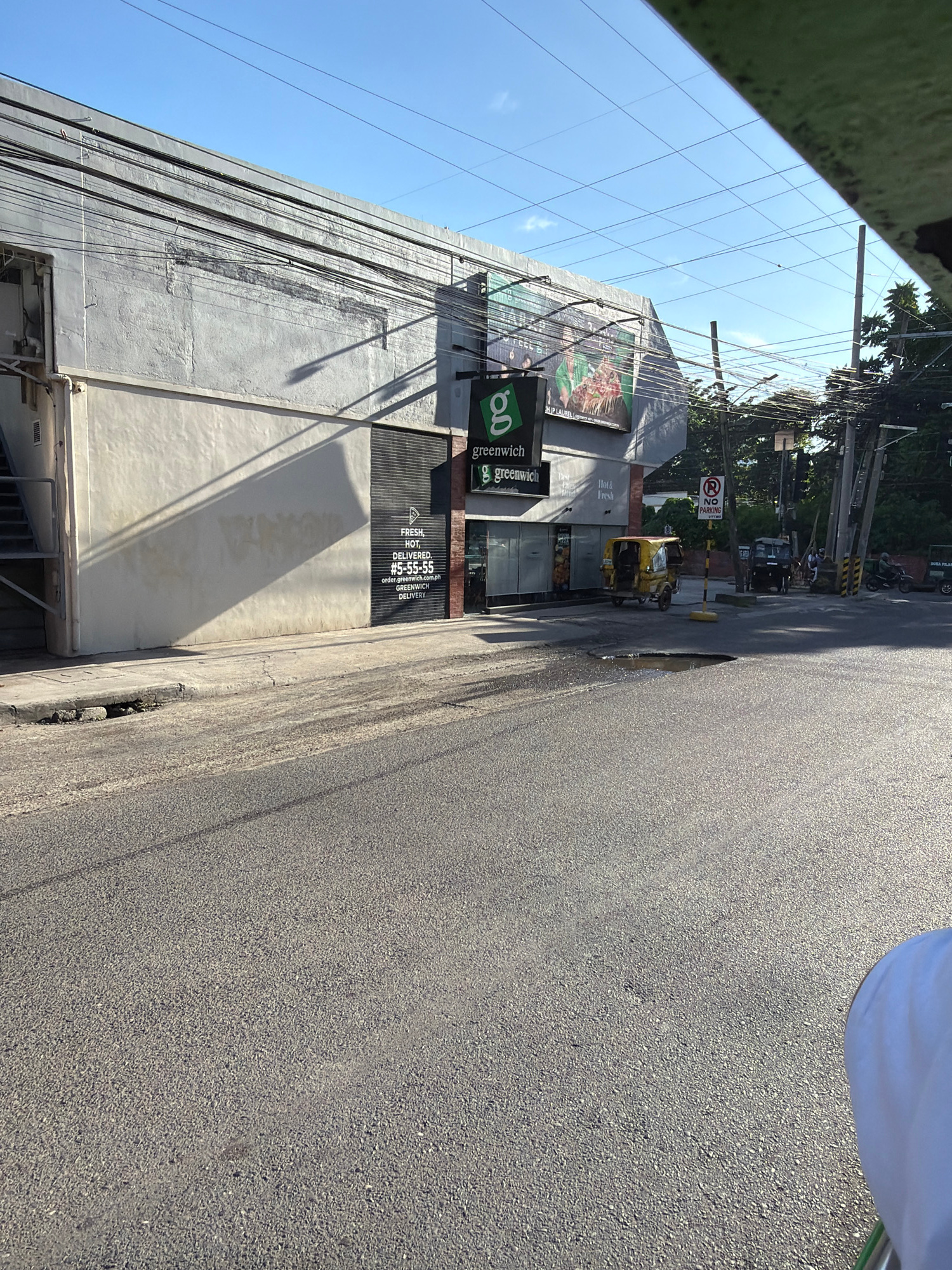 The image captures an exterior view of a Greenwich pizza establishment in Davao City, Philippines, as suggested by the "Greenwich" branding and "DAVAO CITY" text visible on a sign within the frame. The scene unfolds on a sunny day, with a clear blue sky above. The Greenwich storefront features its signature green logo and signage, with an advertisement for hot and fresh pizza. A phone number for delivery, #5-55-55, is also displayed.
Adjacent to the Greenwich establishment, a yellow tricycle, a common mode of transport in the Philippines, is parked. In the background, utility poles and electrical wires crisscross the sky, characteristic of urban landscapes in the region. A "NO PARKING" sign is visible, indicating traffic regulations. The road in the foreground appears to be asphalt and shows signs of wear, including a noticeable pothole filled with water. The overall environment suggests a commercial street in Davao City.