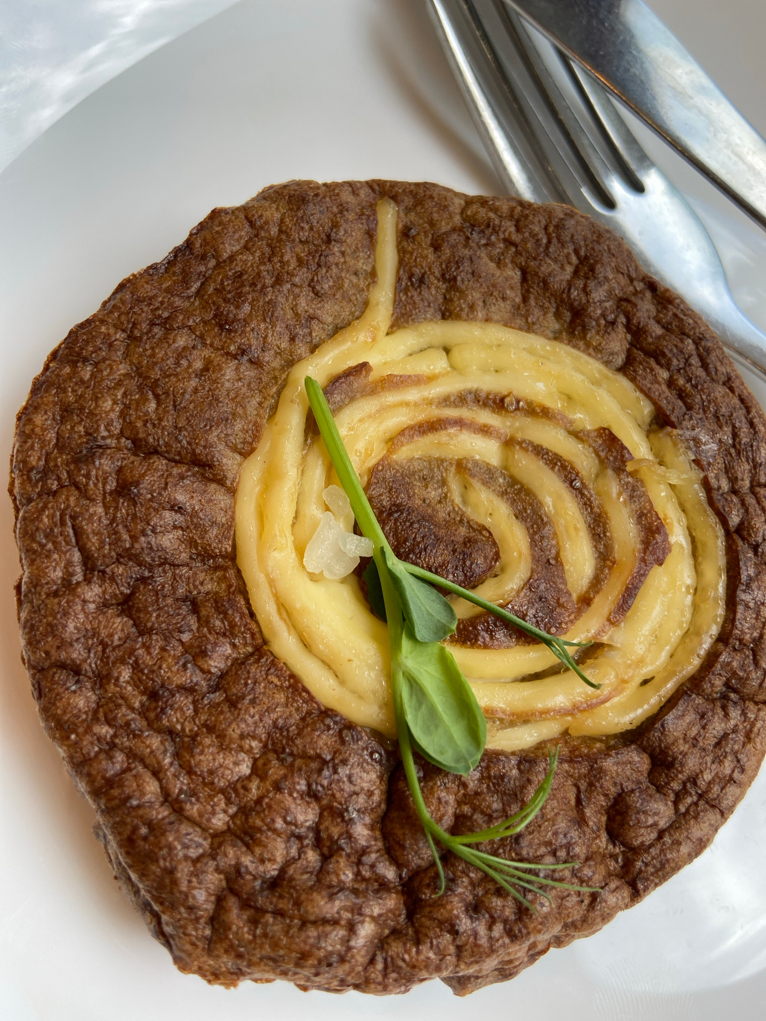 A dark brown, circular dish with a textured, slightly cracked surface is presented on a white plate. A spiral pattern of pale yellow slices, possibly potatoes or cheese, is arranged in the center of the dish. A sprig of green herb, possibly microgreens, with a few tiny white pieces of garnish, is placed on top of the spiral. A fork and knife are visible to the upper right of the dish, positioned on the white plate. The background is blurred, showing a white surface and hints of light. The image captures a close-up, overhead view of the prepared food item. There is no identifiable location in the city of Ufa, Russia.