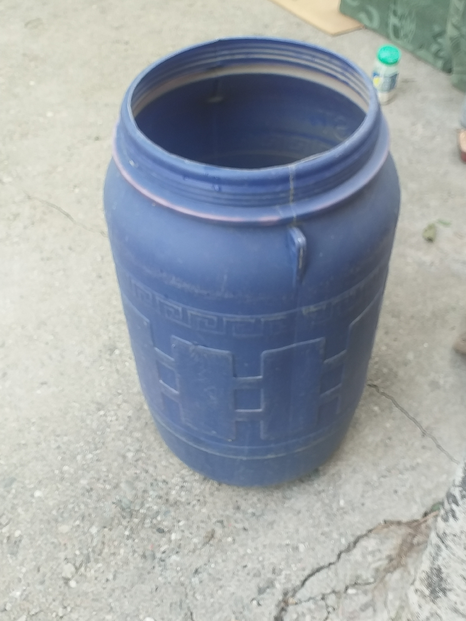 A dark blue, wide-mouthed barrel with decorative rectangular patterns is situated on a cracked concrete surface. The barrel has a thick rim with two distinct ridges. The surface appears to be outdoors, possibly in a yard or alleyway. A small white and green plastic bottle is visible in the upper right background, behind the barrel, suggesting it is not in a pristine or commercial setting. The lighting indicates it is daytime, with shadows cast on the ground. The overall scene is unremarkable and lacks any specific identifying landmarks that would pinpoint a location within Dushanbe, Tajikistan.