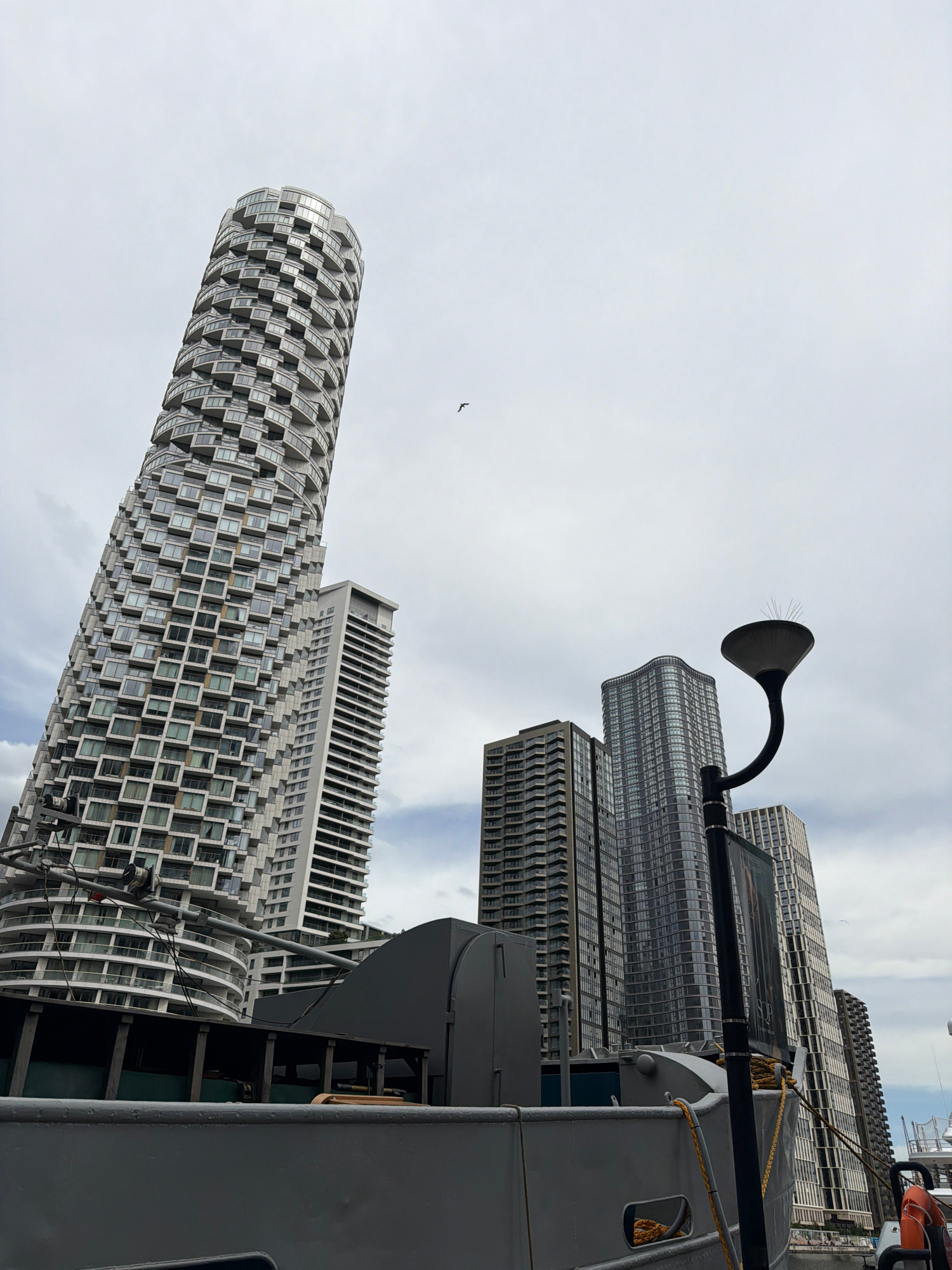 The image shows a view of the South Quay area in Canary Wharf, London, United Kingdom. The foreground is dominated by the deck and side of a grey boat. A lamppost with bird deterrents stands on the right side of the boat. In the background, several modern skyscrapers rise into a cloudy sky. The most prominent building on the left is the distinctive, cylindrical Strata SE1 tower, characterized by its white facade and staggered balconies. Other tall buildings visible include Wardian London and Baltimore Tower, with their glass and metal exteriors. A single bird is visible in flight against the overcast sky.