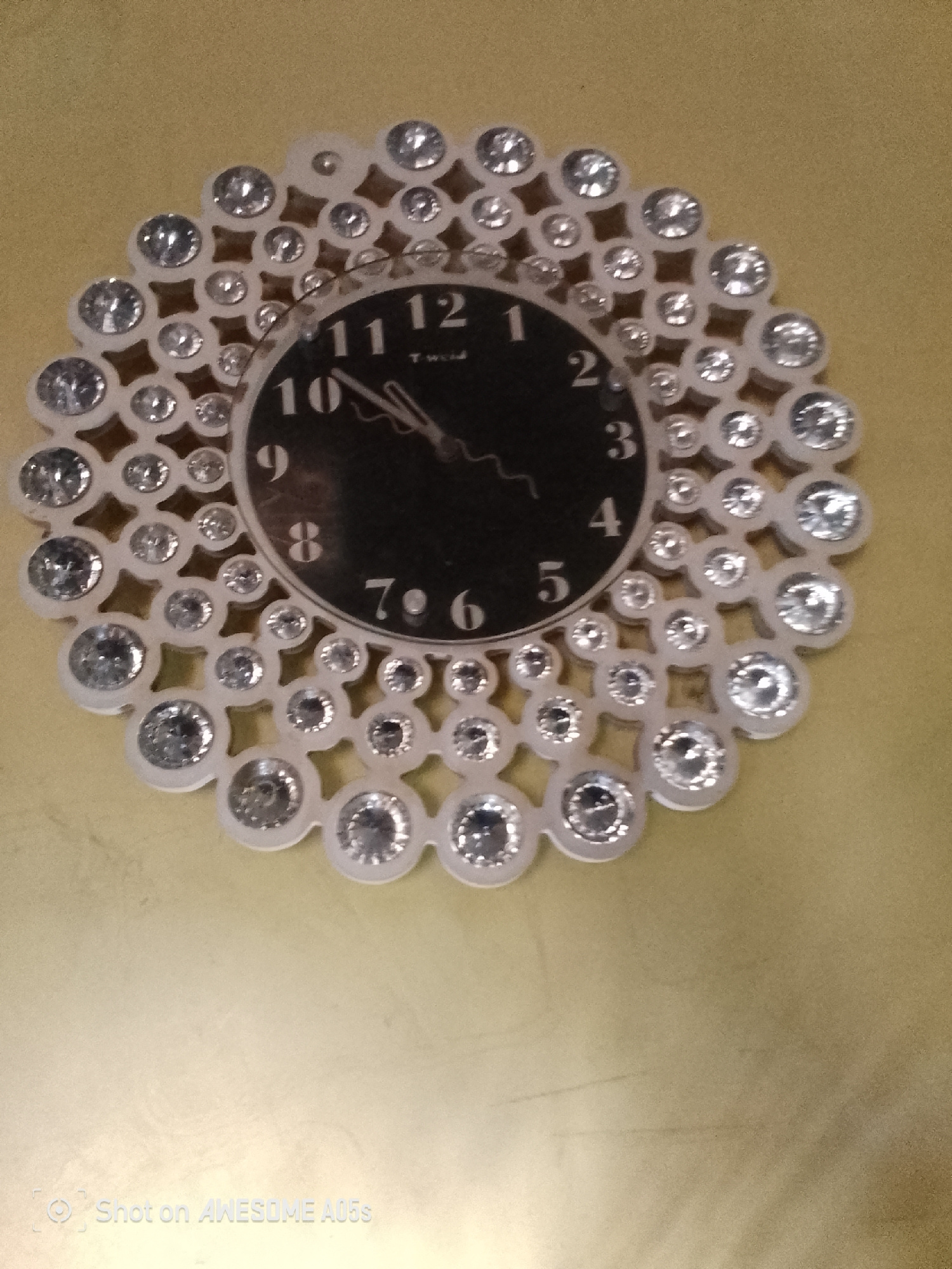 A wall clock with a decorative outer ring is pictured. The clock face is black with white numbers. The outer ring is white and adorned with numerous clear, faceted gemstones. The clock is mounted on a plain, light yellow wall. There is a watermark at the bottom of the image that reads "Shot on AWESOME A05s". The image does not contain any information about the city of Jauro Yinu, Nigeria.