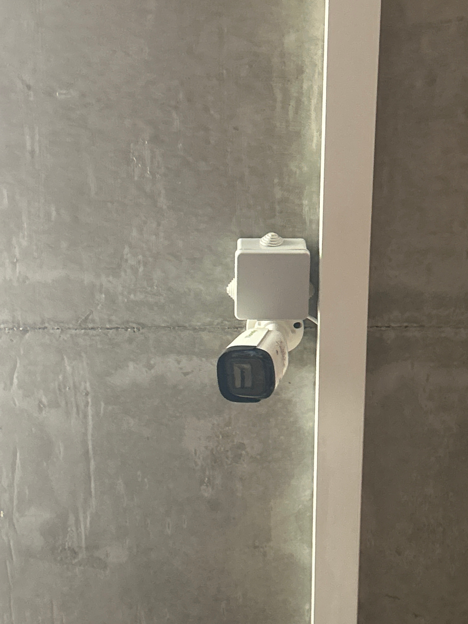A security camera is mounted on a textured grey concrete ceiling. The camera is white with a black lens. A white junction box is attached above the camera. A vertical white beam is to the right of the camera, and a line is visible horizontally across the concrete wall below the camera. The image appears to be taken indoors. This image was captured in Tbilisi, Georgia.