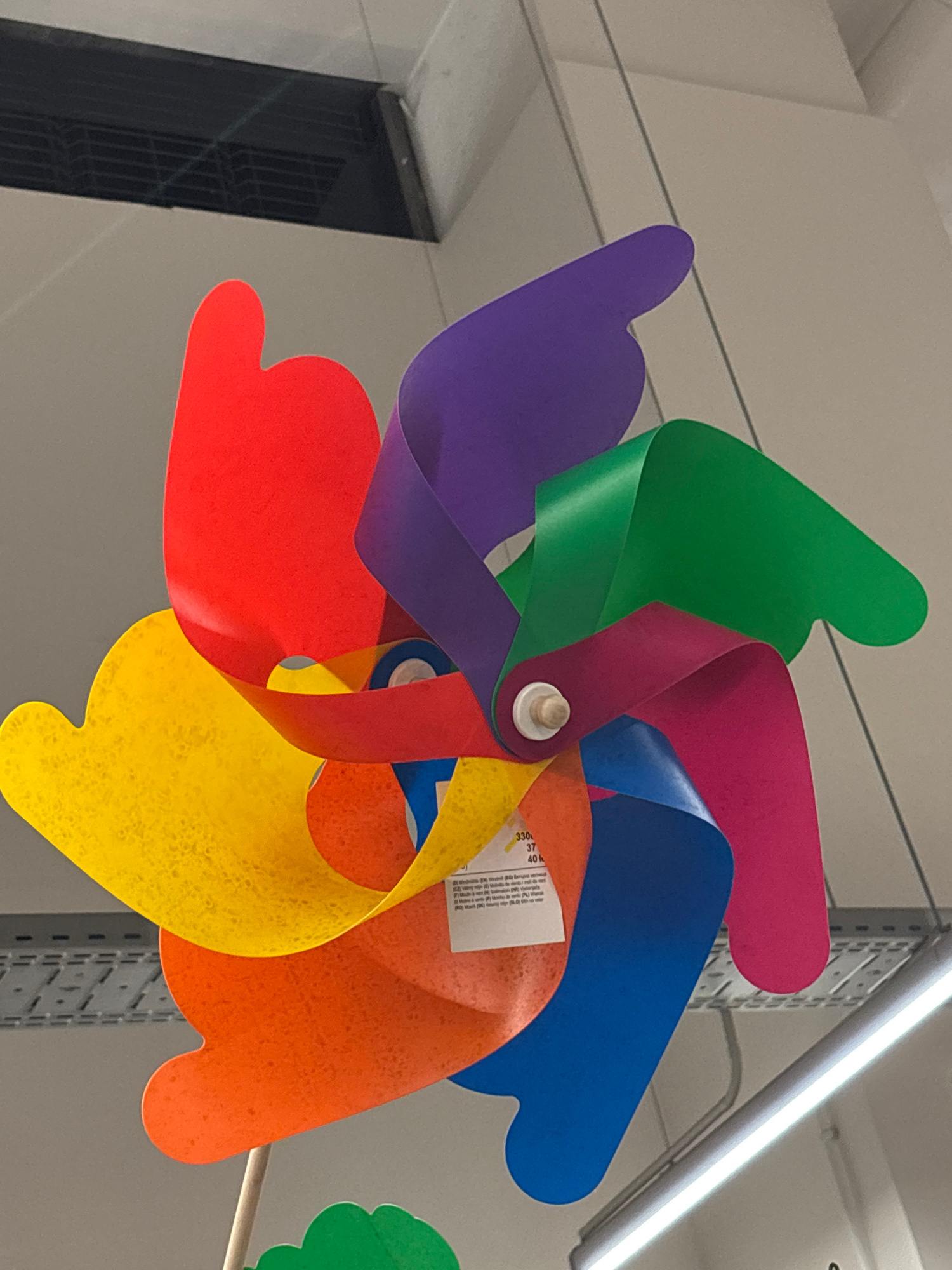 A rainbow-colored pinwheel is displayed against a neutral background. The pinwheel has six blades, each a different color of the rainbow: red, orange, yellow, green, purple, and blue. The blades are curved and appear to be made of plastic. A small white label is attached to one of the blades. The pinwheel is mounted on a thin wooden stick. The background suggests an indoor setting, possibly a store or a market, with a black ventilation grate visible in the upper left corner and a fluorescent light fixture running horizontally in the lower right. The overall impression is that of a colorful, playful object. The location is stated to be in the city of Varna, Bulgaria.