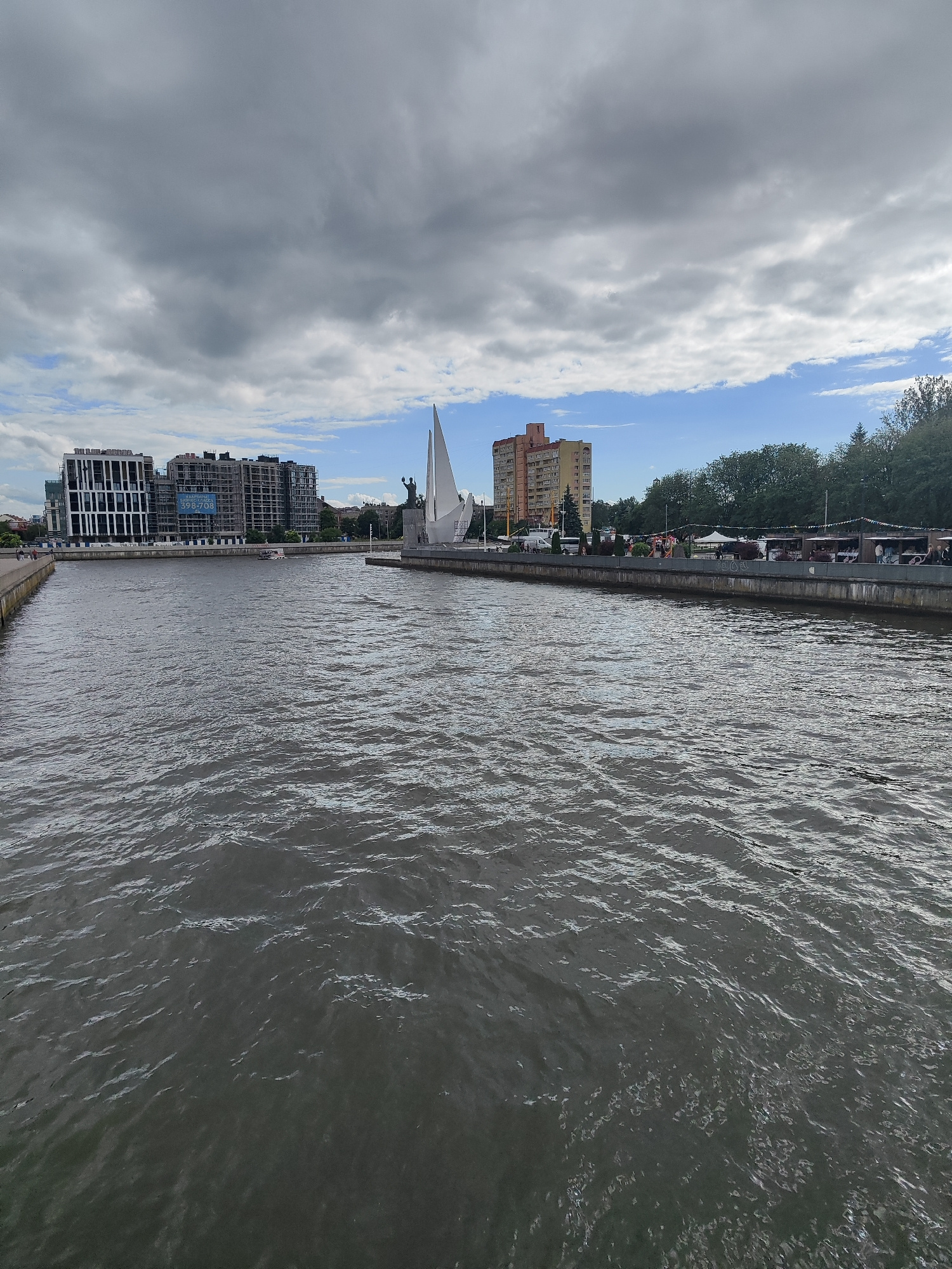The image captures a scene along the Pregolya River in Kaliningrad, Russia, likely near the Fischdorf (Fish Village) area. In the foreground, the dark, rippling water of the river dominates the lower half of the frame. On the left bank, a concrete embankment is visible, leading towards a modern building complex under construction, characterized by its sleek, dark-paneled facade and exposed concrete structures. On the right bank, a more developed promenade is evident, lined with trees and several market stalls or kiosks, suggesting an active public space. In the middle ground, a distinctive white sail-shaped sculpture stands prominently, accompanied by a bronze statue of a figure on a pedestal. A small boat is seen moving across the water, and buildings of various architectural styles, including a multi-story residential building with a distinctive orange facade, rise behind the trees. The sky above is a mix of heavy gray clouds and patches of blue, indicating a partly cloudy day. The overall impression is a blend of urban development, historical landmarks, and natural elements along the riverfront.