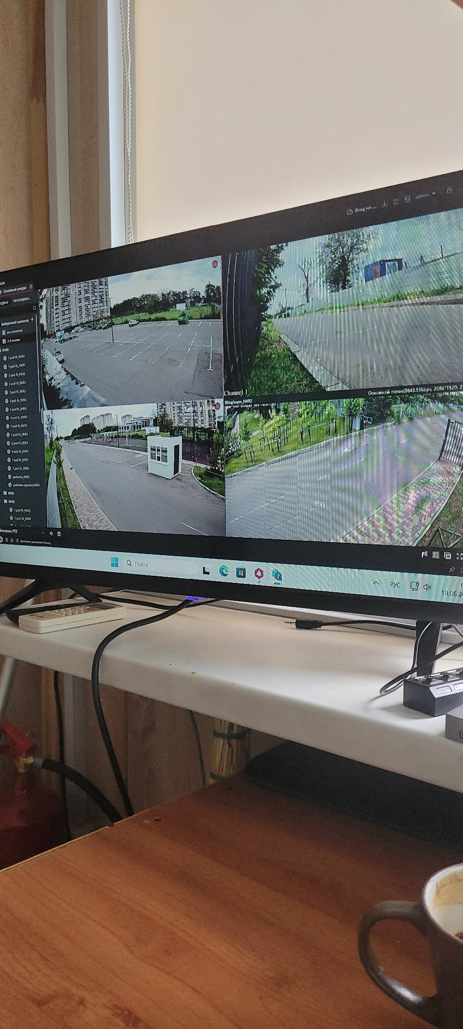 The image shows a computer monitor displaying a multi-camera feed, likely from a surveillance system. The monitor is positioned vertically and is black. The screen displays four different camera views simultaneously. The top two views appear to be of an outdoor area with a road, grass, and some trees. One of these top views has text overlay that reads "Основной поток (2643.55kbps, 268811520," and "Channel 1 Shlagbaym NVR2". The bottom two views show what appears to be a parking lot with marked spaces and some buildings in the background. One of these bottom views has a small white structure in the middle of the lot.
On the left side of the monitor, there is a vertical menu with icons and text, including "РУС" which indicates the language is Russian. There is also a search bar with the text "Q Поиск". At the bottom of the screen, there is a list of camera channels labeled "NVR1", "NVR2", etc., and further text that appears to be camera names or identifiers. A date, "19.06.20," is visible.
The monitor is placed on a white surface, likely a desk or table, next to a wooden surface. A black cable runs from the monitor and connects to something off-camera. A dark-colored mug with a lighter interior is visible on the wooden surface, suggesting a possible workspace environment. The lighting is somewhat dim, suggesting it could be daytime, but the artificial light from the monitor is prominent. There are no people visible in the image. The overall scene depicts a monitoring station for a security or surveillance system, likely in Krasnodar, Russia, given the Cyrillic text.