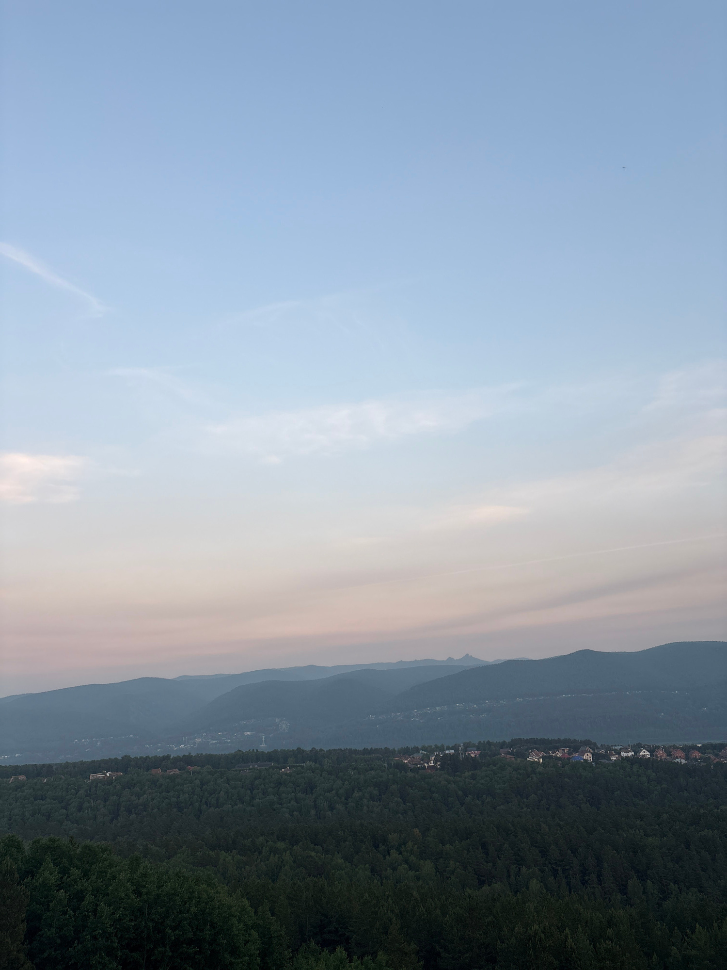 This image captures a panoramic view from a high vantage point, likely overlooking the city of Krasnoyarsk, Russia, at dusk. The sky transitions from a clear, pale blue at the top to soft hues of pink and orange closer to the horizon, indicating the end of the day. wispy clouds are scattered across the sky.
Below the sky, a range of silhouetted mountains forms the backdrop. The slopes of these mountains are covered in dense, dark green forest. In the middle ground, nestled among the trees, a cluster of houses with red and brown roofs is visible, suggesting a suburban or semi-rural settlement. The trees in the foreground are also a deep green, with some lighter green foliage suggesting deciduous trees.
The overall impression is one of natural beauty and tranquility, with the human settlement appearing small and integrated within the vast landscape. There are no people, animals, or specific actions depicted in the scene. The image is a static, observational record of the landscape at a particular time of day.