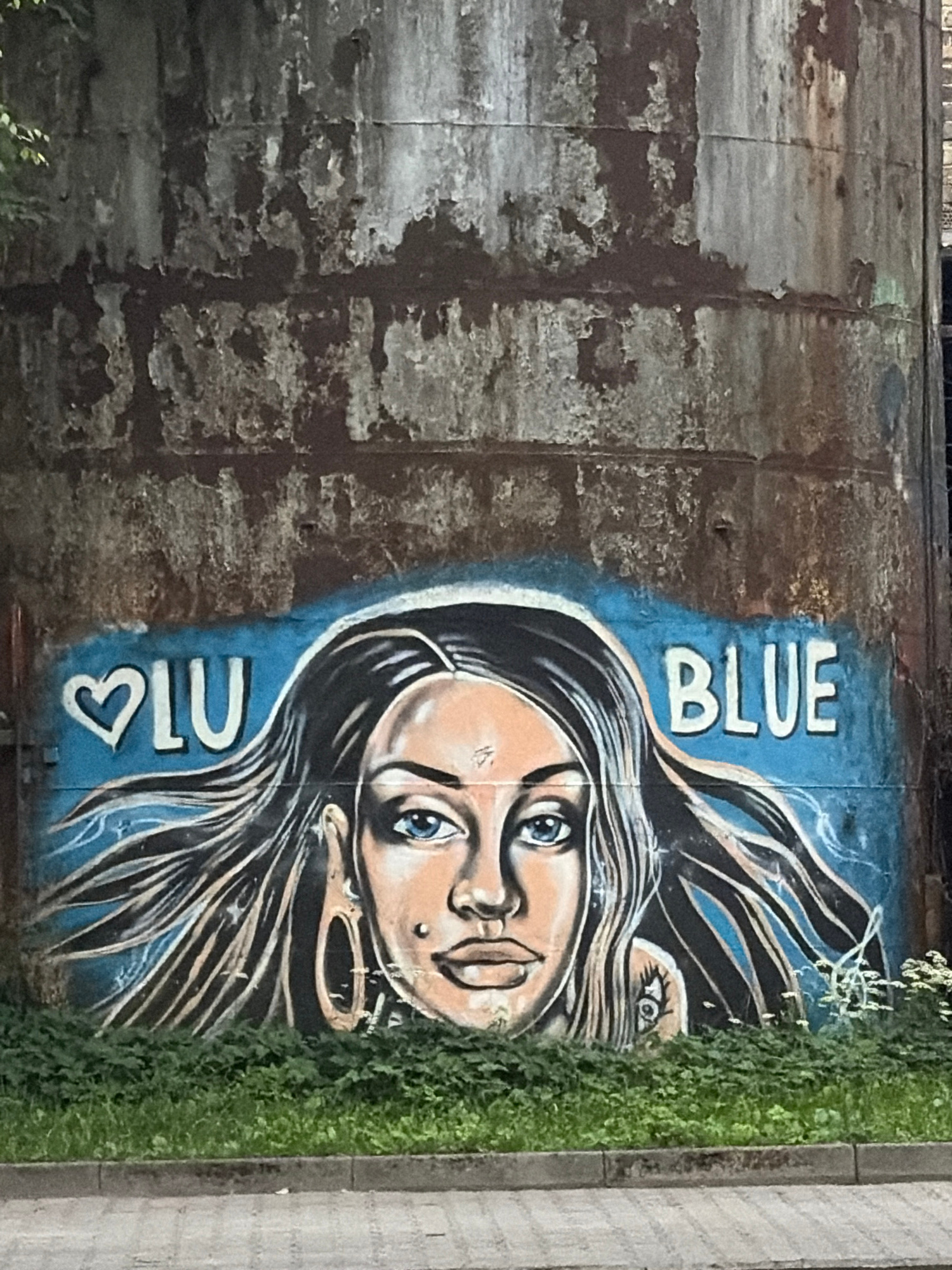 A graffiti mural is painted on a tall, rusty, cylindrical structure. The mural depicts the upper portion of a woman's face with long, flowing black hair against a blue background. To the left of the face, the word "ILU" is written, with a heart symbol replacing the "I". To the right of the face, the word "BLUE" is painted. The cylindrical structure is weathered and shows signs of rust and peeling paint. Green vegetation grows at the base of the structure. The foreground shows a paved surface and a curb. The graffiti is stylized, with bold lines and bright colors contrasting with the weathered texture of the structure.