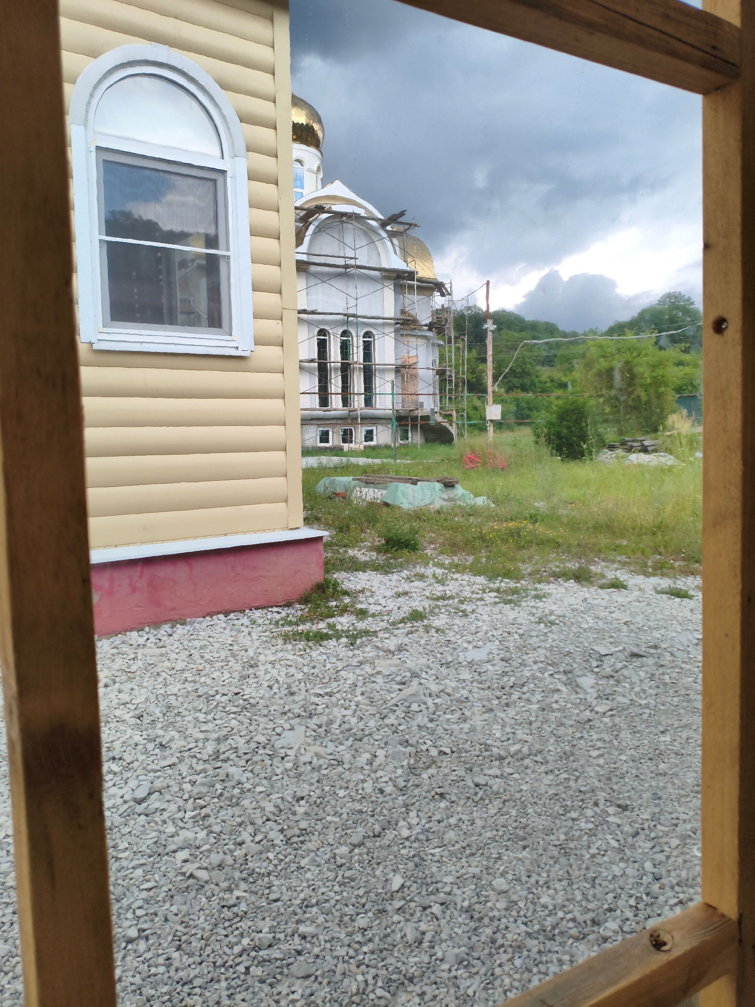 The image is a view through a wooden frame of a construction site in Vozrozhdeniye, Russia. In the foreground, to the left, is a section of a light-colored, horizontal wooden-paneled building with a red-painted foundation. To the right of this building, the main subject is a large white building under construction, possibly a church, with scaffolding and construction materials around it. This building features arched windows and a golden dome. Behind the construction site, lush green trees and a hilly landscape are visible under a cloudy, grey sky. The ground in the foreground is covered with light-colored gravel.