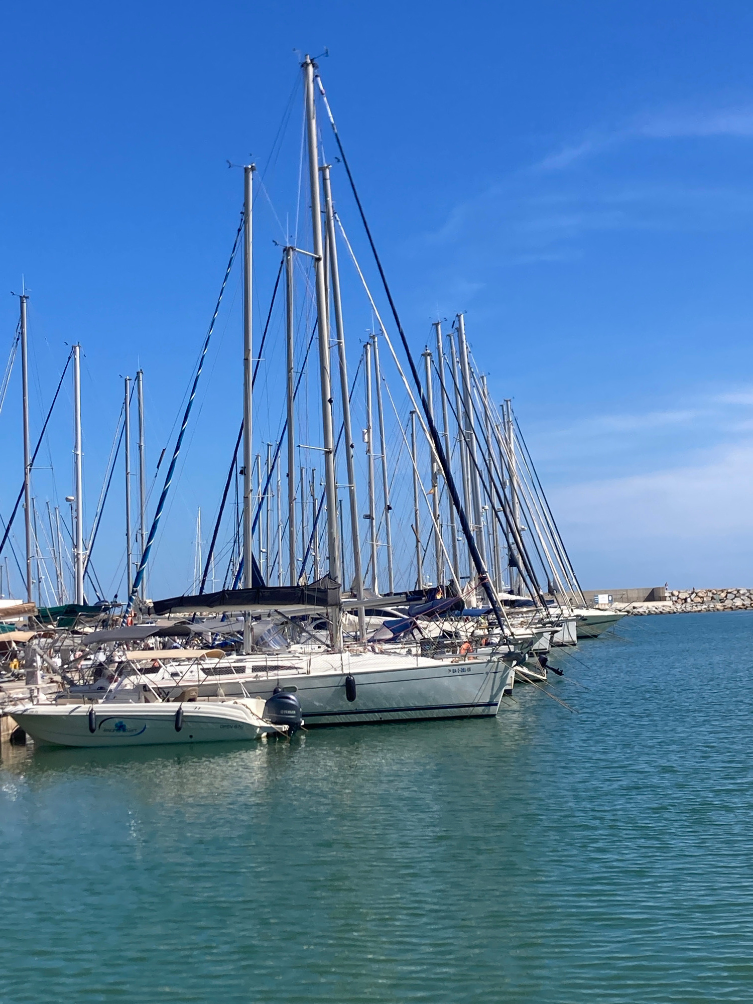 The image shows a marina with numerous sailboats docked on a clear, blue day. The boats are lined up parallel to the viewer, with their masts reaching high into the sky. The water is a calm, turquoise color. In the background, a stone breakwater is visible, and beyond that, a clear blue sky with a few wispy clouds. One of the boats in the foreground is a smaller white motorboat with a Yamaha outboard motor and a logo on its side. Another sailboat in the foreground has the registration number "71 BA-2-261-08" visible on its hull. The location appears to be Port De La Ginesta, Spain, suggested by the style of the boats and the characteristic Mediterranean blue sky. There are no people clearly visible in the image.