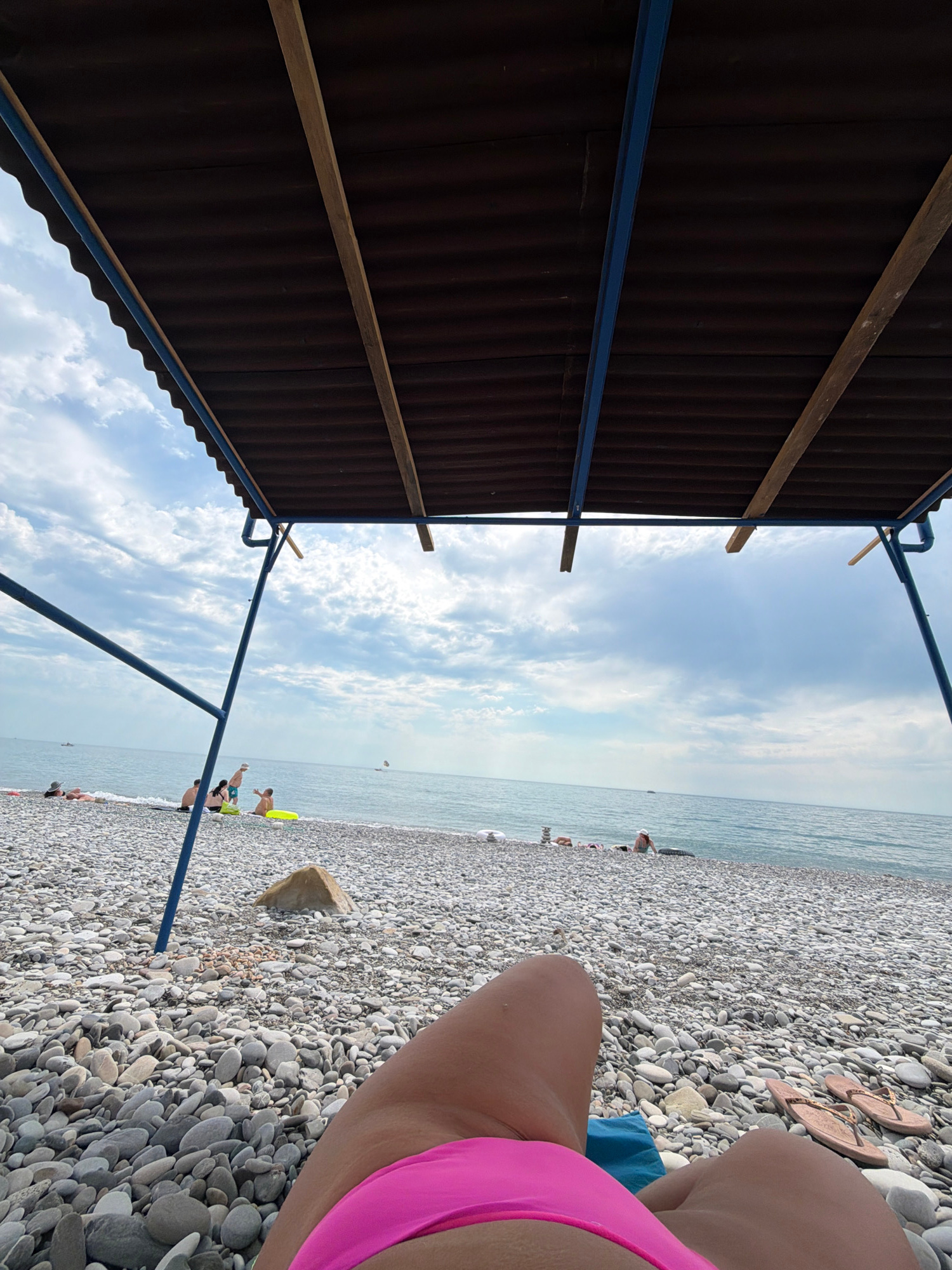 A low-angle, vertically oriented shot captures a beach scene in Sochi, Russia. In the foreground, a person's legs and hip, wearing a bright pink bikini, are visible from the side, resting on a pebbled beach. Scattered on the pebbles are a pair of flip-flops and a blue towel. The beach is covered in smooth, gray and white pebbles, stretching towards the sea.
In the middle ground, several people are scattered along the shoreline. Some are sitting or lying on the beach, while others appear to be walking or standing near the water's edge. One person is visible in the water, and another is wearing a yellow float.
The background features the calm, grayish-blue sea meeting the sky under a partly cloudy, bright sky. A dark, corrugated awning supported by blue metal poles and wooden beams dominates the left side of the frame, casting a shadow over a portion of the beach. The sun appears to be high in the sky, suggesting it's daytime. The overall mood is relaxed and typical of a beach day. No visible text is present in the image.