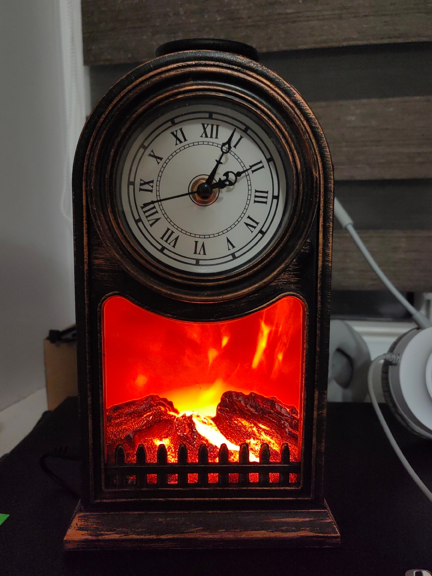The image shows a decorative electric fireplace clock. The clock face features Roman numerals and has a white background with black markings. The hour and minute hands are black. The fireplace part of the object is illuminated with a vibrant orange and red glow, simulating flames. The frame of the object appears to be made of a dark, possibly metallic material with an antique finish. The object is placed on a dark surface, and in the background, a white cord and white headphones are partially visible. The overall scene is dimly lit, with the exception of the bright glow from the artificial fireplace. No specific location within the city of Hrodna, Belarus can be determined from the provided image.