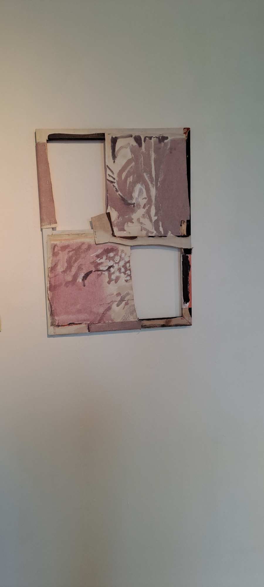 The image shows a framed abstract artwork hanging on a plain, light-colored wall. The artwork is composed of several pieces of paper or canvas, layered and adhered to a backing. The colors are predominantly muted pinks, purples, and beige, with some dark lines and shapes. The frame appears to be made of wood, with a dark border. The artwork is positioned centrally in the frame, with white space visible through cutouts in the paper layers. The lighting suggests an indoor setting, possibly a home or gallery. There are no people or specific objects that would indicate a particular location within Castlebar, Ireland. The overall impression is of a contemporary art piece.
