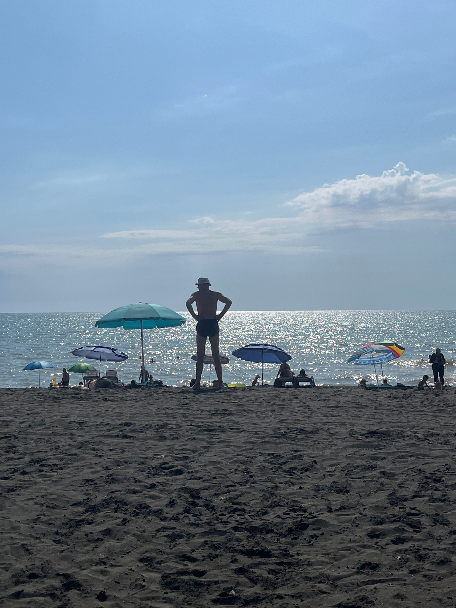 The image depicts a sunny day at Ureki Beach in Georgia, known for its black magnetic sand. In the foreground, dark, textured sand stretches across the frame, with footprints and ripples indicating recent activity. The middle ground shows a line of beach umbrellas and loungers, casting shadows on the sand. Several people are visible, some relaxing under the umbrellas, while others are standing or walking near the water's edge. A prominent silhouette of a man wearing a hat and shorts stands in the center, facing the sea with his hands on his hips.
The background is dominated by the Black Sea, its surface shimmering with sunlight. The water appears calm, with small waves lapping the shore. A hazy horizon separates the sea from a pale blue sky, which is dotted with a few wispy clouds. The overall atmosphere is one of leisure and relaxation on a warm summer day.